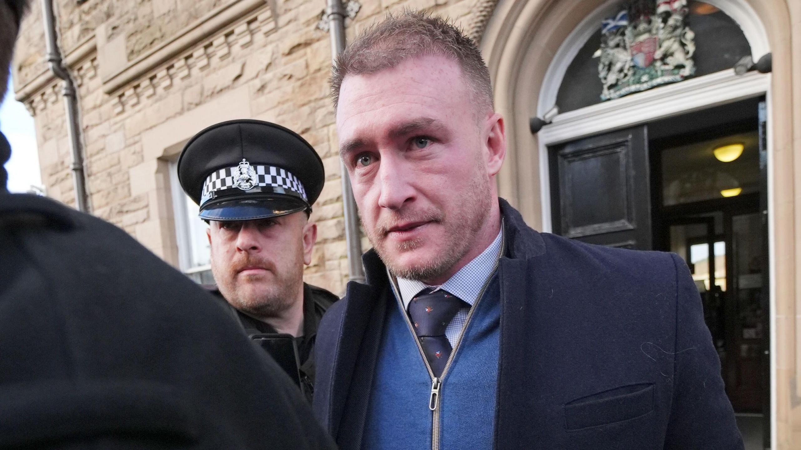 A man with a stubbly beard and short hair heads out of a court building wearing a zip up top and shirt and tie while accompanied by a police officer