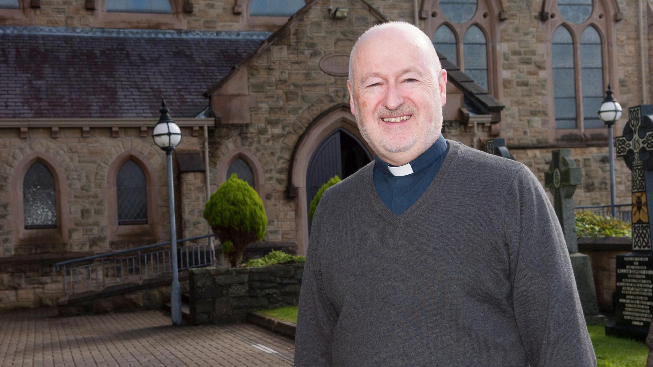 Fr John Murray stands outside in front of a stone-brick church. He wears a grey jumper over a navy shirt with a white priest collar. 
