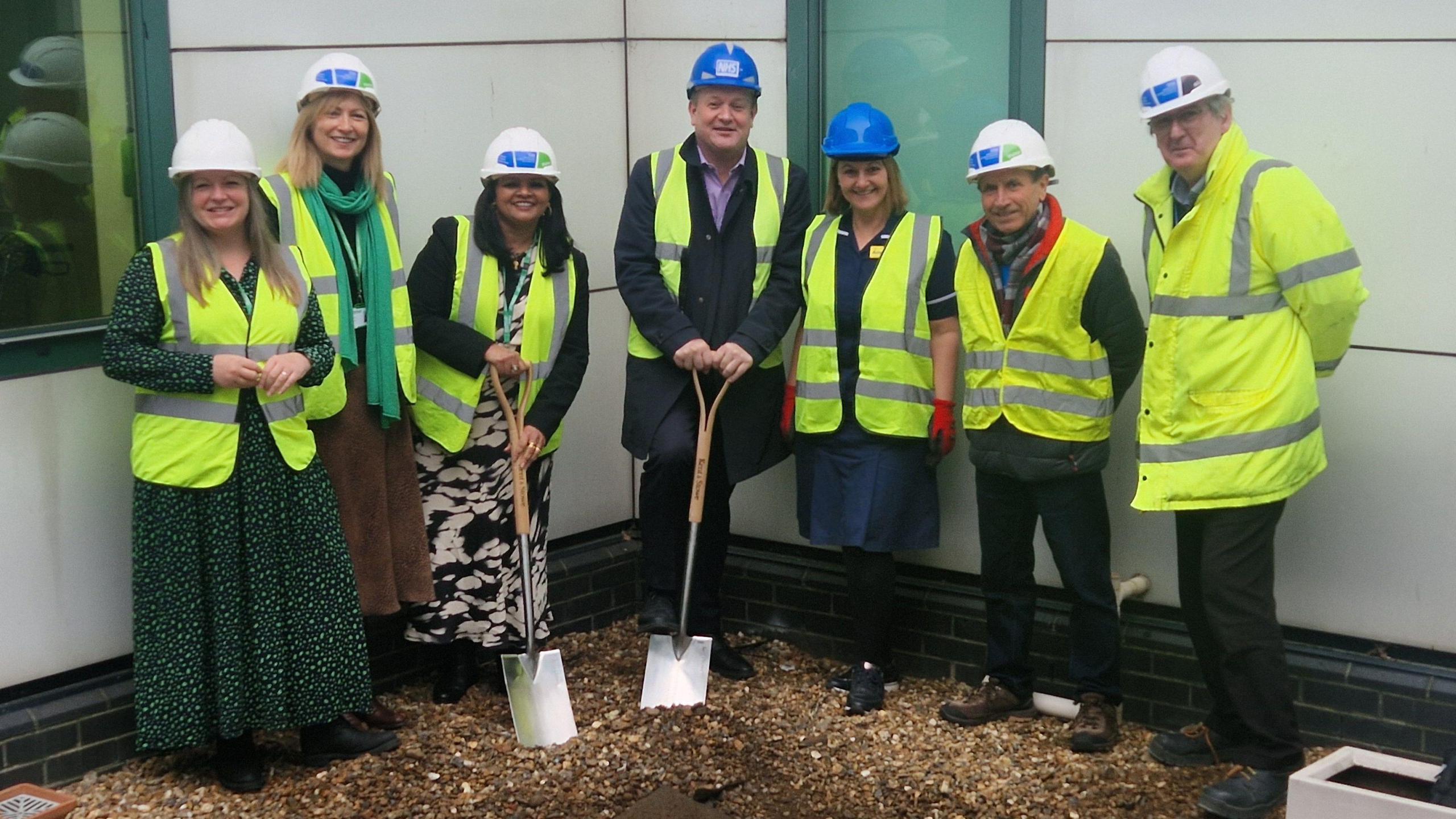 Seven people from the Royal Surrey NHS Foundation Trust pictured at the Ashford Hospital in Surrey, where ground has broken on the construction of a new cancer centre. All seven people are wearing high-viz and hard hats, and a woman third from left and a man fourth from left are holding spades.