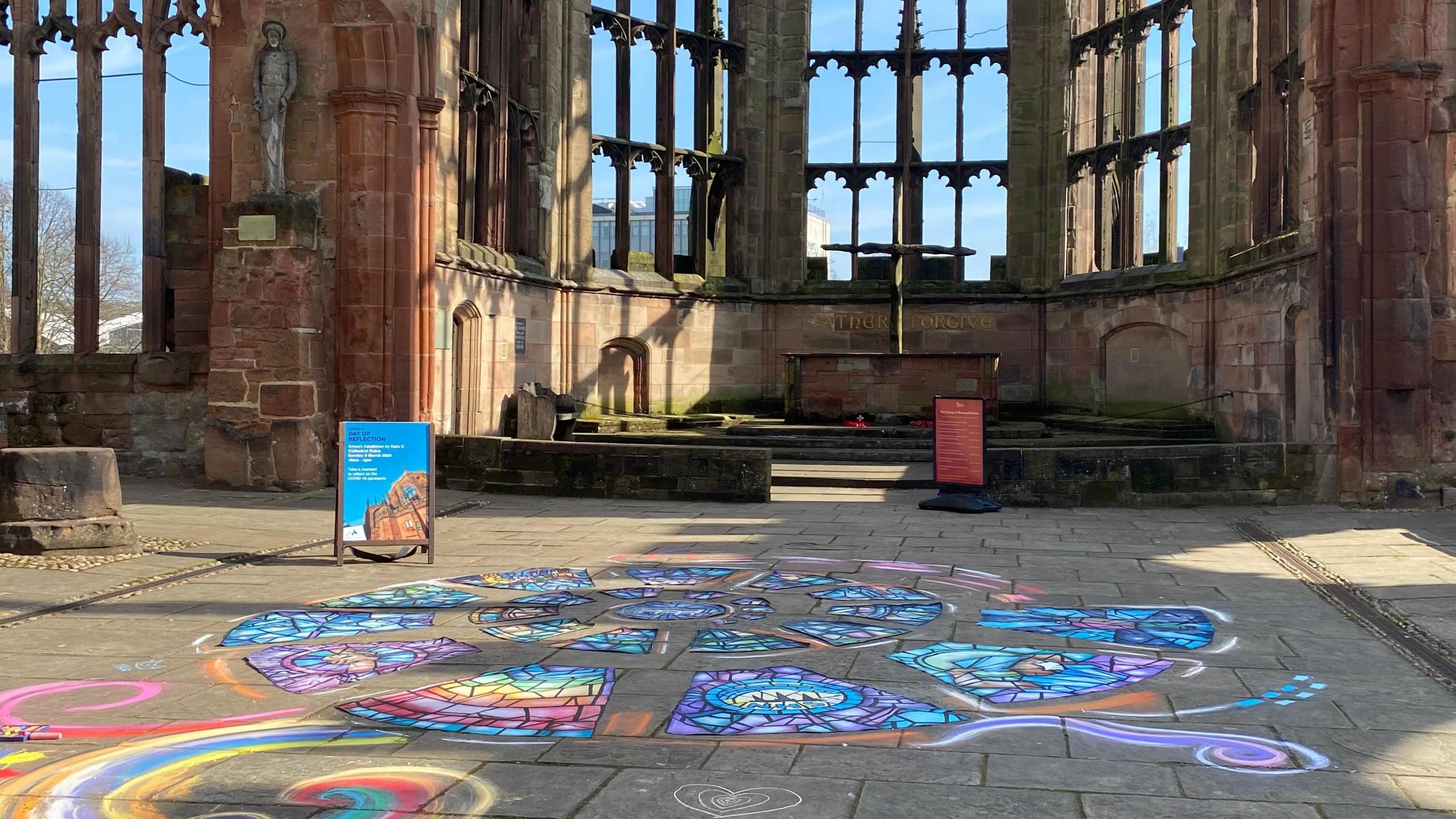 A colourful, spiralling artwork resembling a stained glass window has been created on paving slabs on the ground. The window-less frames of Coventry Cathedral's stone structure are visible behind, with blue skies.