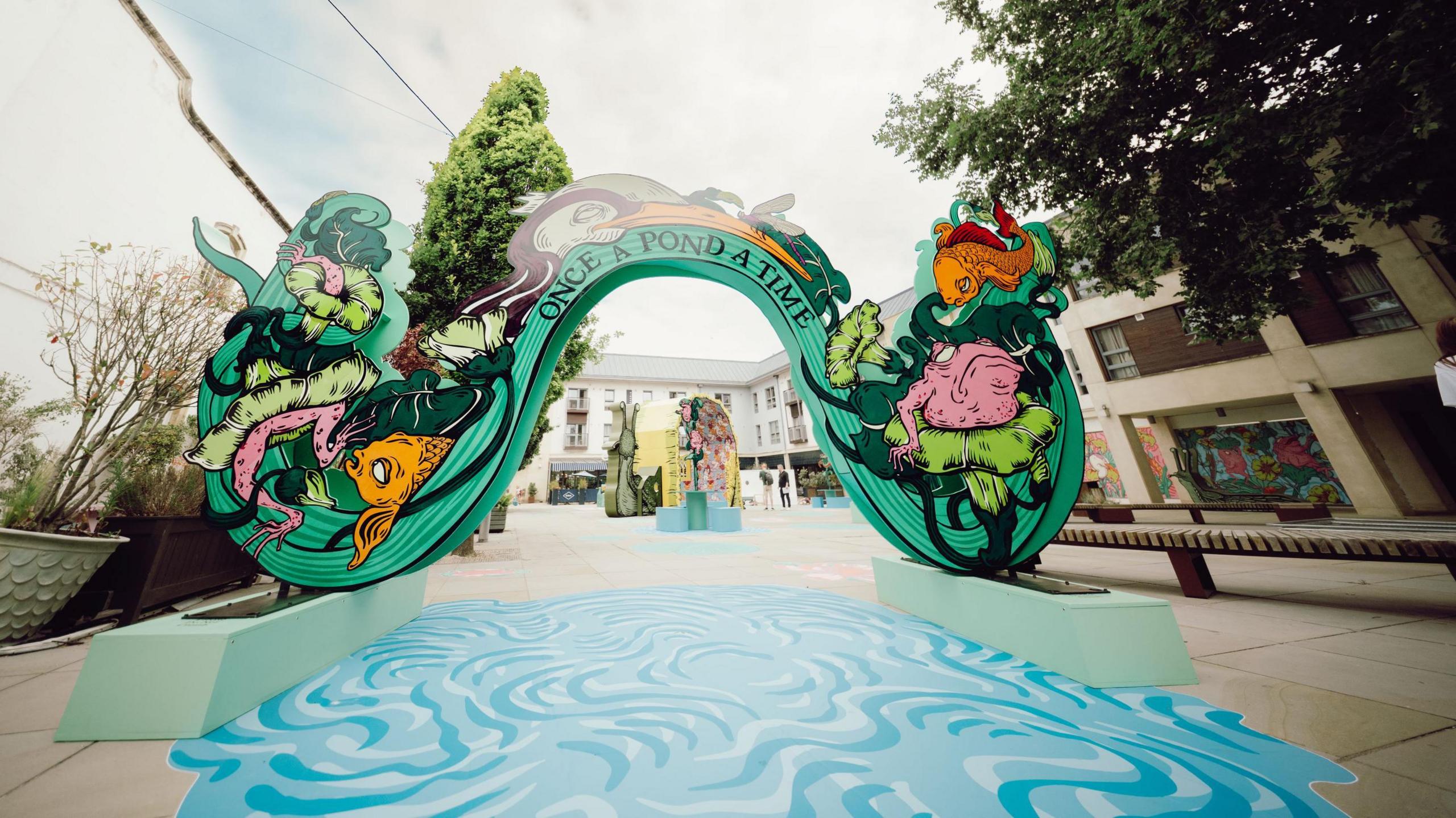 An art installation called "Once A Pond a Time" is seen in Quakers Friars in Bristol. It has a blue area painted on the floor to look like water, and an arch with colourful characters painted on it, including an orange fish and a pink frog.