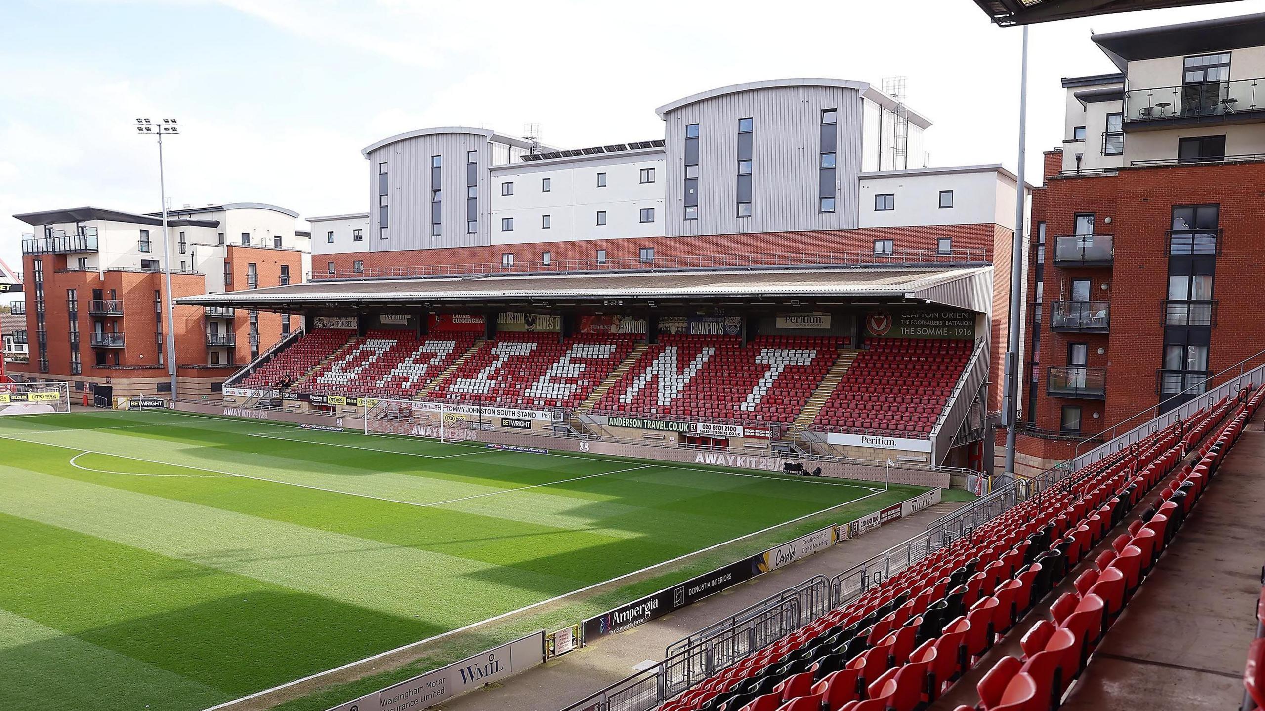 A view from the side of Leyton Orient's stadium, a football stand with red seats with Orient painted in white .