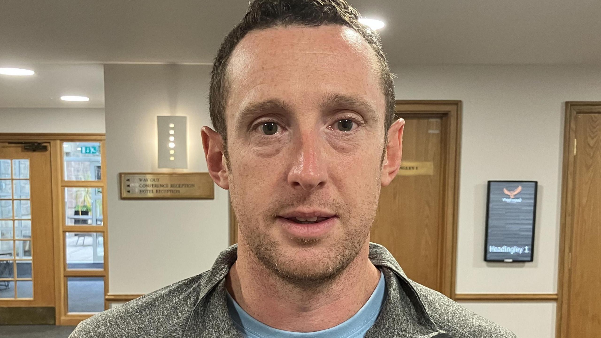 A man with short brown hair and brown eyes wearing a light blue t shirt and a grey cycling jacket. He is standing in an office building with signs on the wall behind him and doors with signs on them.