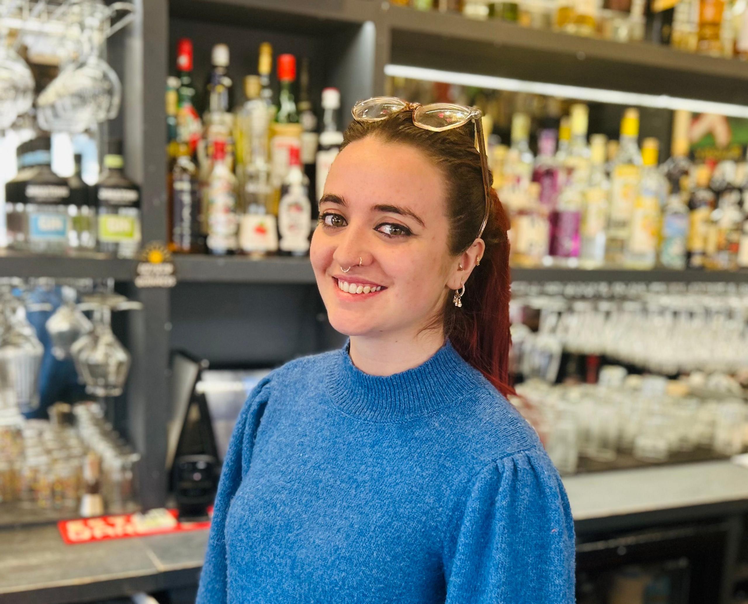 A mid shot of a woman in a blue jumper. She is standing behind a bar and bottles are blurred in the background. She has red hair which is tied up in a high ponytail and her glass are on her head.