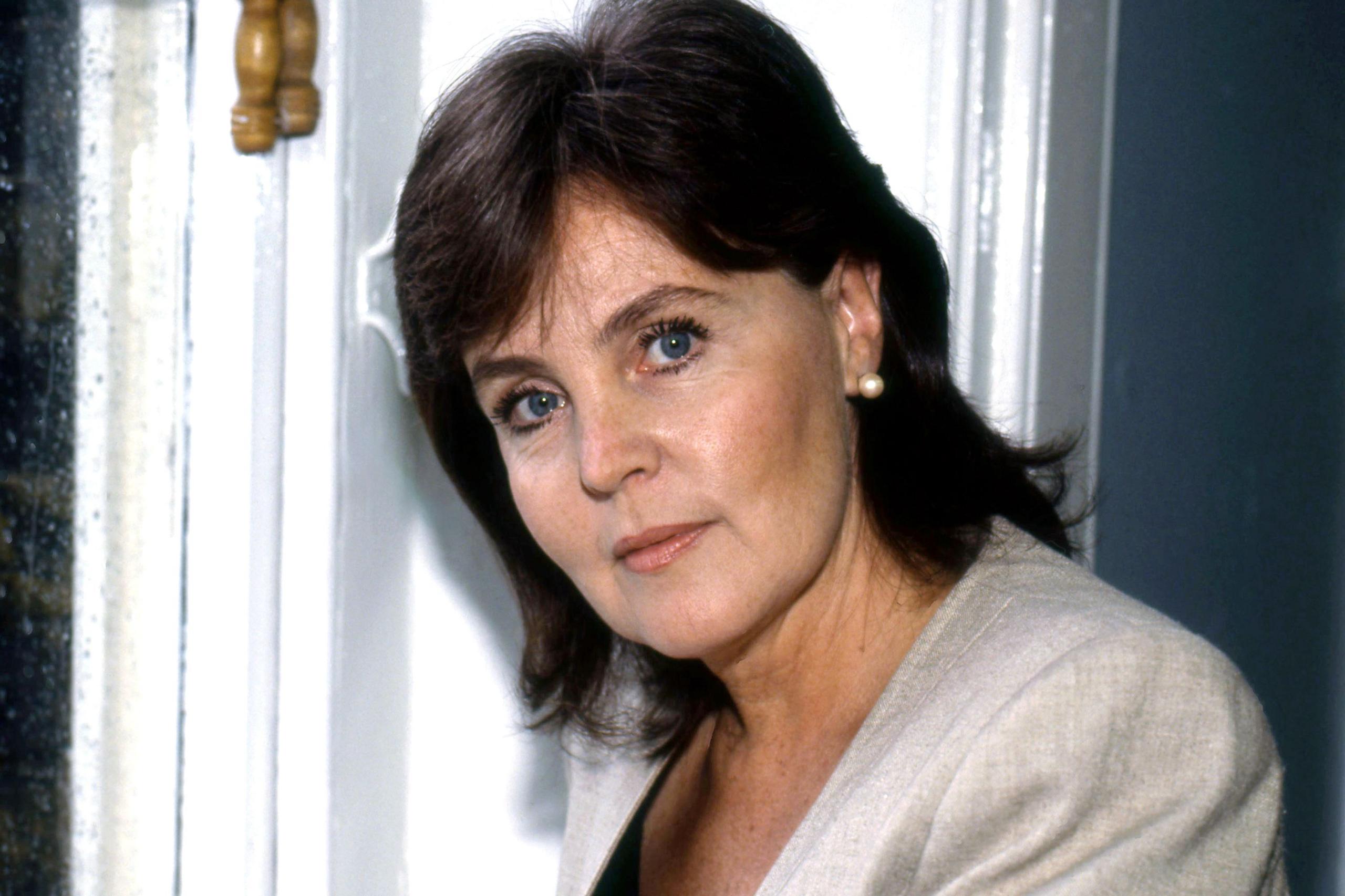 Pauline Collins ka veshur një xhaketë gri të çelët mbi një bluzë të errët, të shoqëruar me një vath të vetëm me perla. Sfondi përfshin një kornizë dere të bardhë dhe një pjesë të një dritareje me një shul druri, duke sugjeruar një ambient të brendshëm me dritë natyrale që depërton.