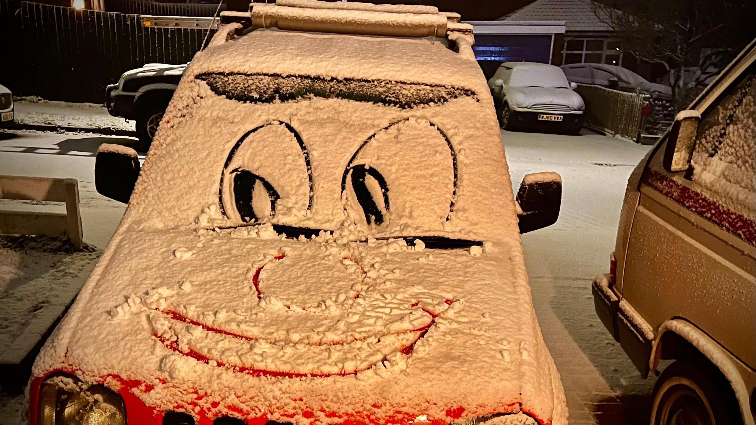 A car with snow on it, someone has drawn a face on it so shows eyes and a big smile. There is snow on the ground and on other cars. 