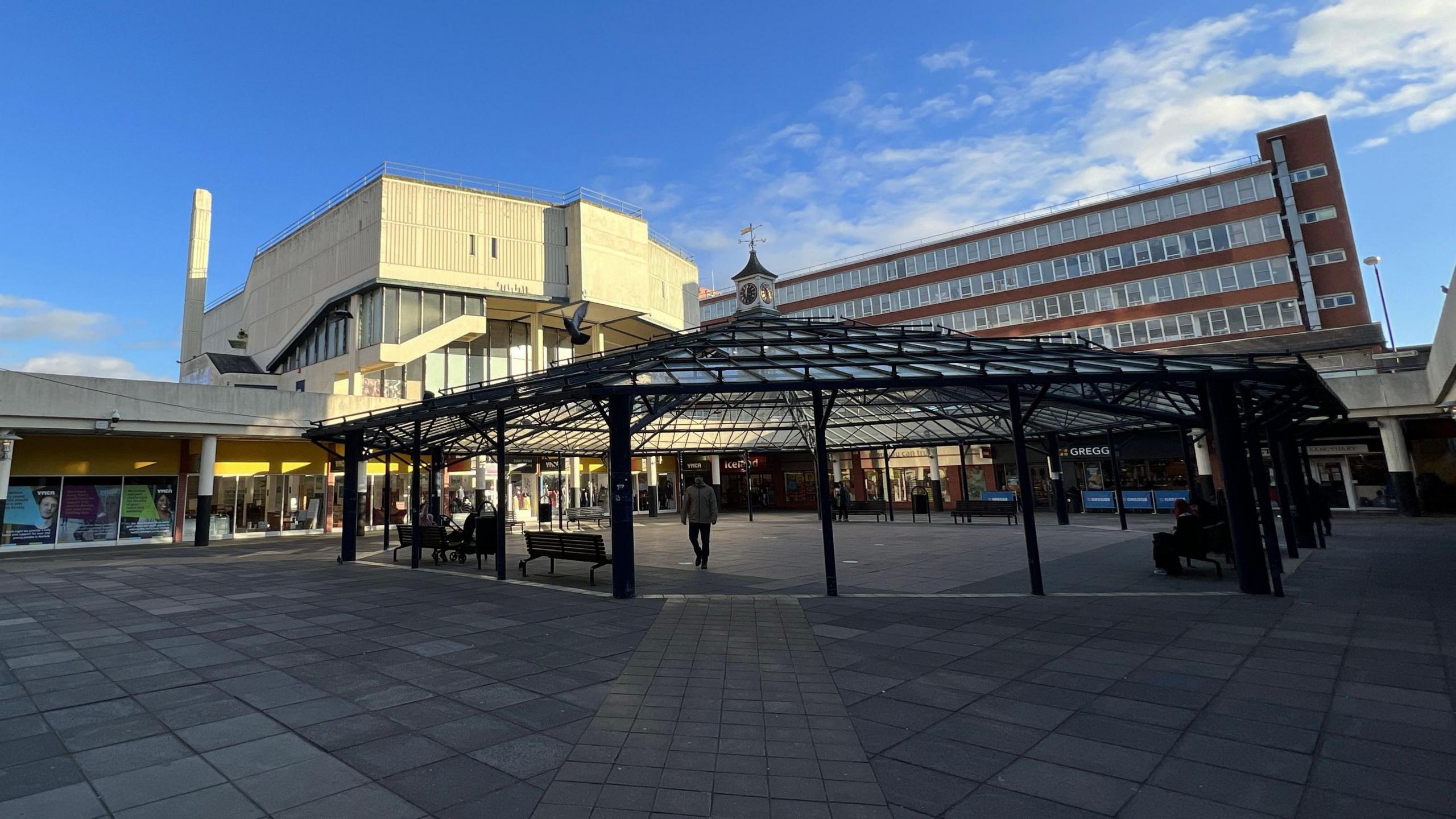 A wide picture of the central precinct of Anglia Square. The middle part is covered by an octagonal metal framework with shops surrounding it. Further afield we can see the old cinema - a large cream building - and an office block.