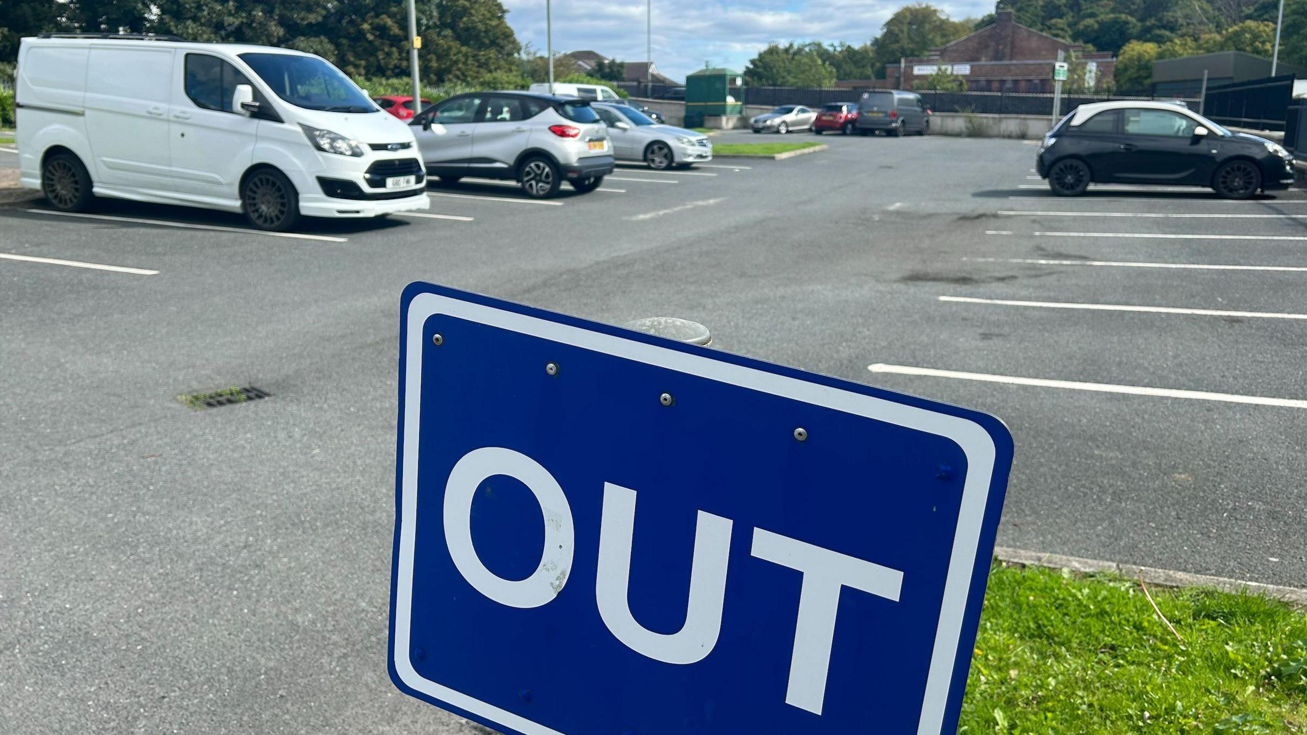 A blue sign with the word 'out' in capital letters on it in the foreground. The Bowl car park has a van and several cars parked in its bays in amongst plenty of empty spaces.