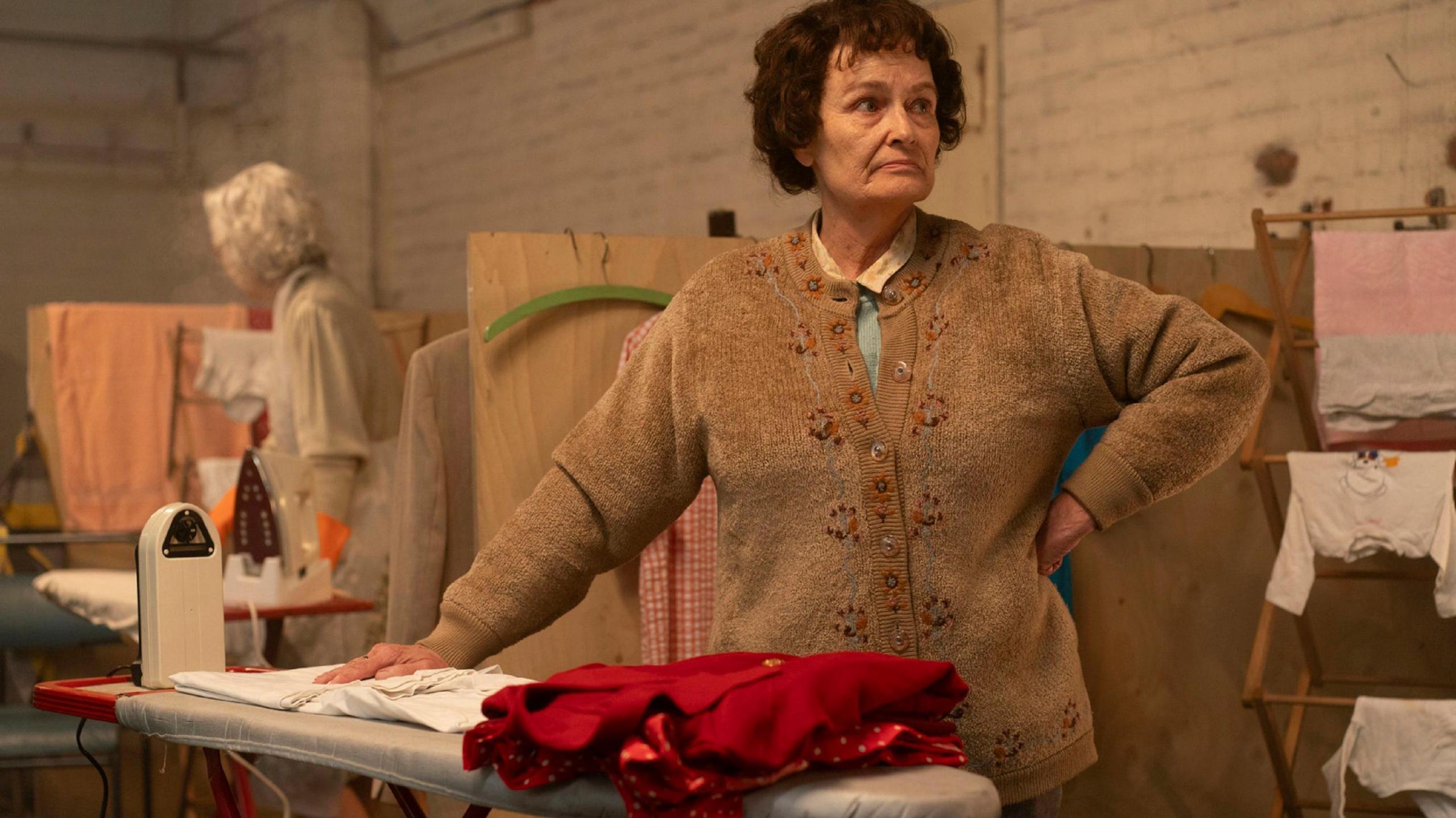 Ella Ramsden, played by Estrid Barton, standing by an ironing board with items of clothing; she's an older woman in a brown cardigan