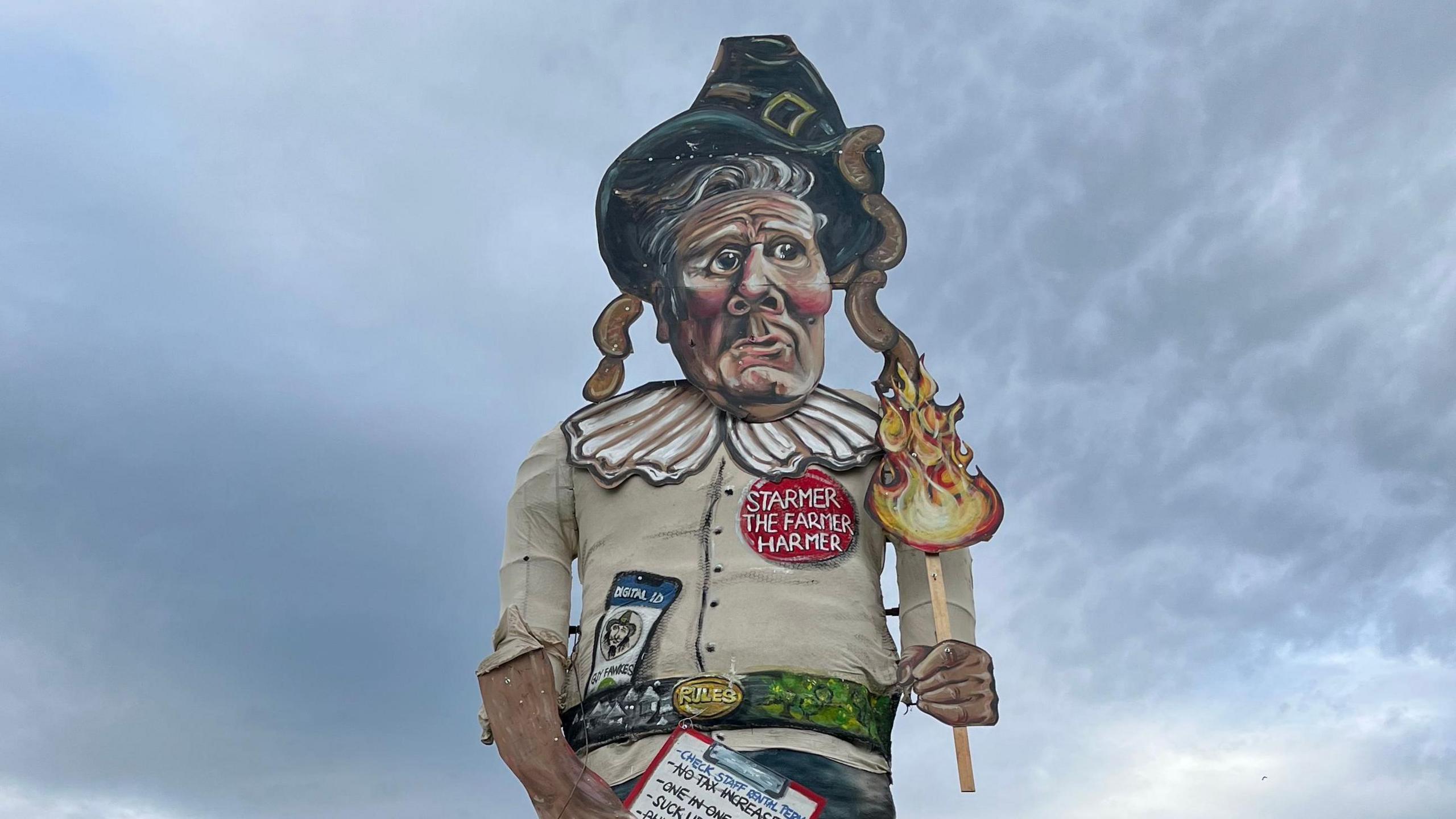 A large effigy of Sir Keir Starmer. The caricature has a Guy Fawkes-style black hat and ruff, is carrying a burning torch, is wearing a red badge that reads 'Starmer the Farmer Harmer' and has an ID badge labeled 'digital ID' on its belt.
