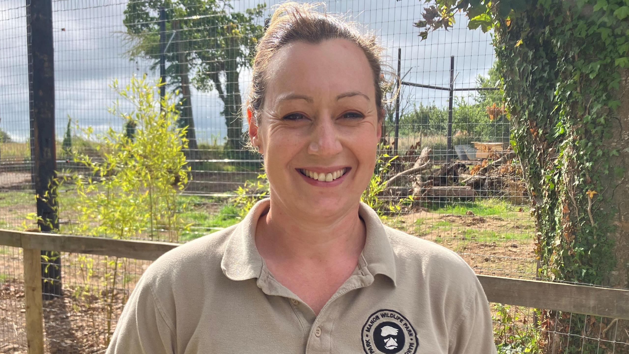 Head keeper at Manor Wildlife Park, Kim Wilkins. She has brown hair pulled back in a ponytail, she wears a light brown polo shirt with a Manor wildlife park badge and smiles showing her teeth. A large enclosure can be seen in the background, along with trees and dried grass.