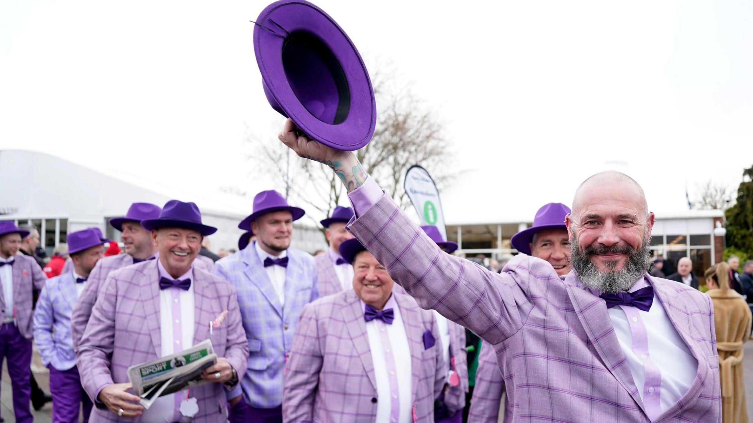 A large group of men wearing purple checked blazers and dark purple dickie bows and hats arrive at the course, one carrying a newspaper and one raising his hat in the air. 