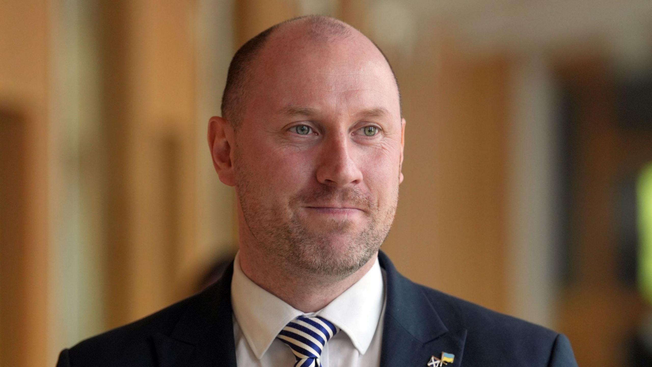 Neil Gray standing in a corridor at the Scottish Parliament building with a smile on his face. He is wearing a dark jacket, a blue and white tie and white shirt.