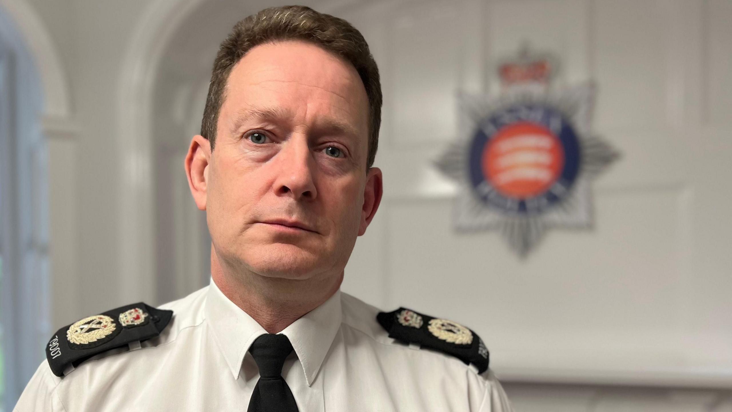 Ben-Julian Harrington, who has short brown hair, staring at the camera with a serious expression. He is wearing a white shirt with a black tie and black epaulettes, which have a golden crest and crown on them. In the background is the blue, red and silver Essex Police crest.