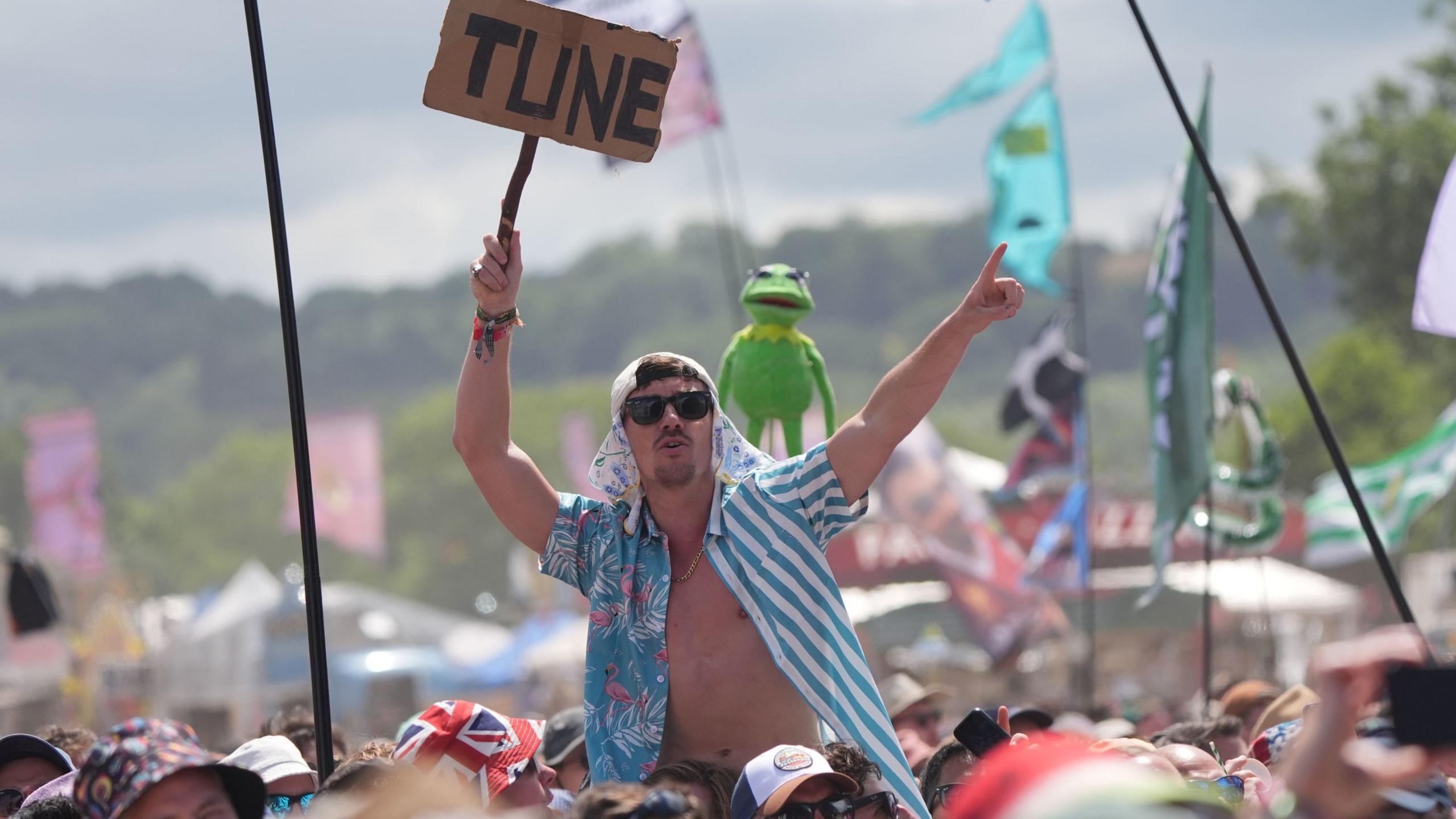 A man wearing an open shirt - blue and white stripes on one side, and blue and flamingo patterned on the other, sits on someone's shoulders in the crowd and holds up a sign saying TUNE. There are crowds all around him and flags being held up