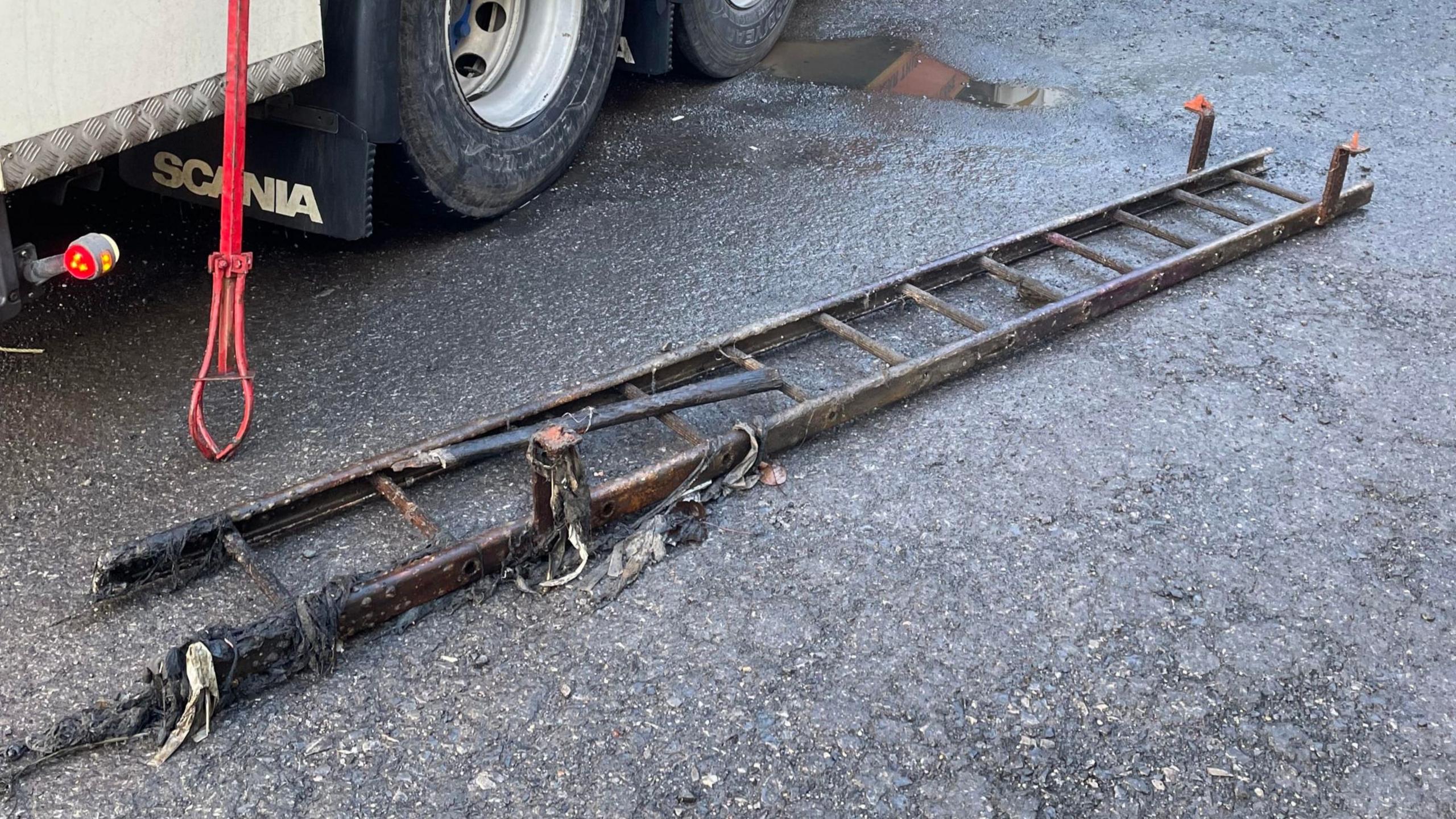 A long metal ladder covered in blackened debris lies on a road. A truck is parked in the background. 