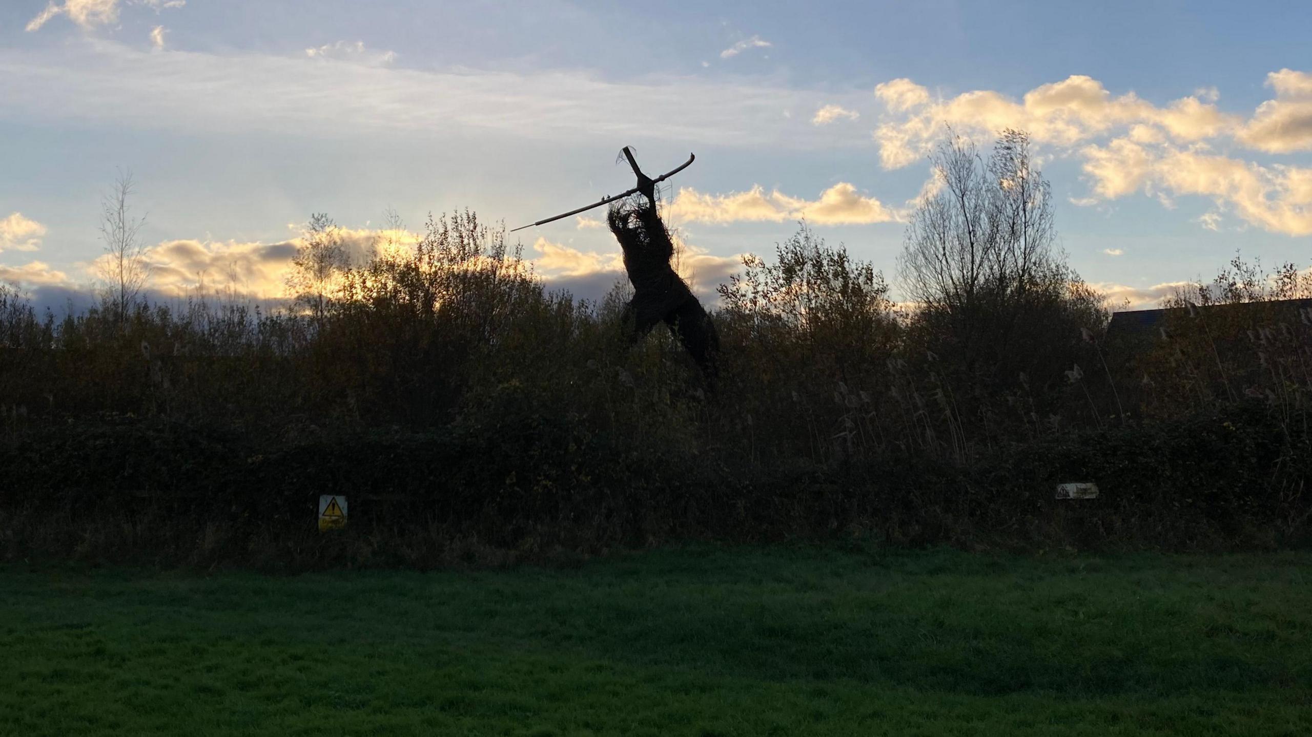 A shot of the Willow Man in the distance, with a field in the foreground while the winter son sets. The structure's top half has worn away and only the frame can be seen, while the waist down appears intact.