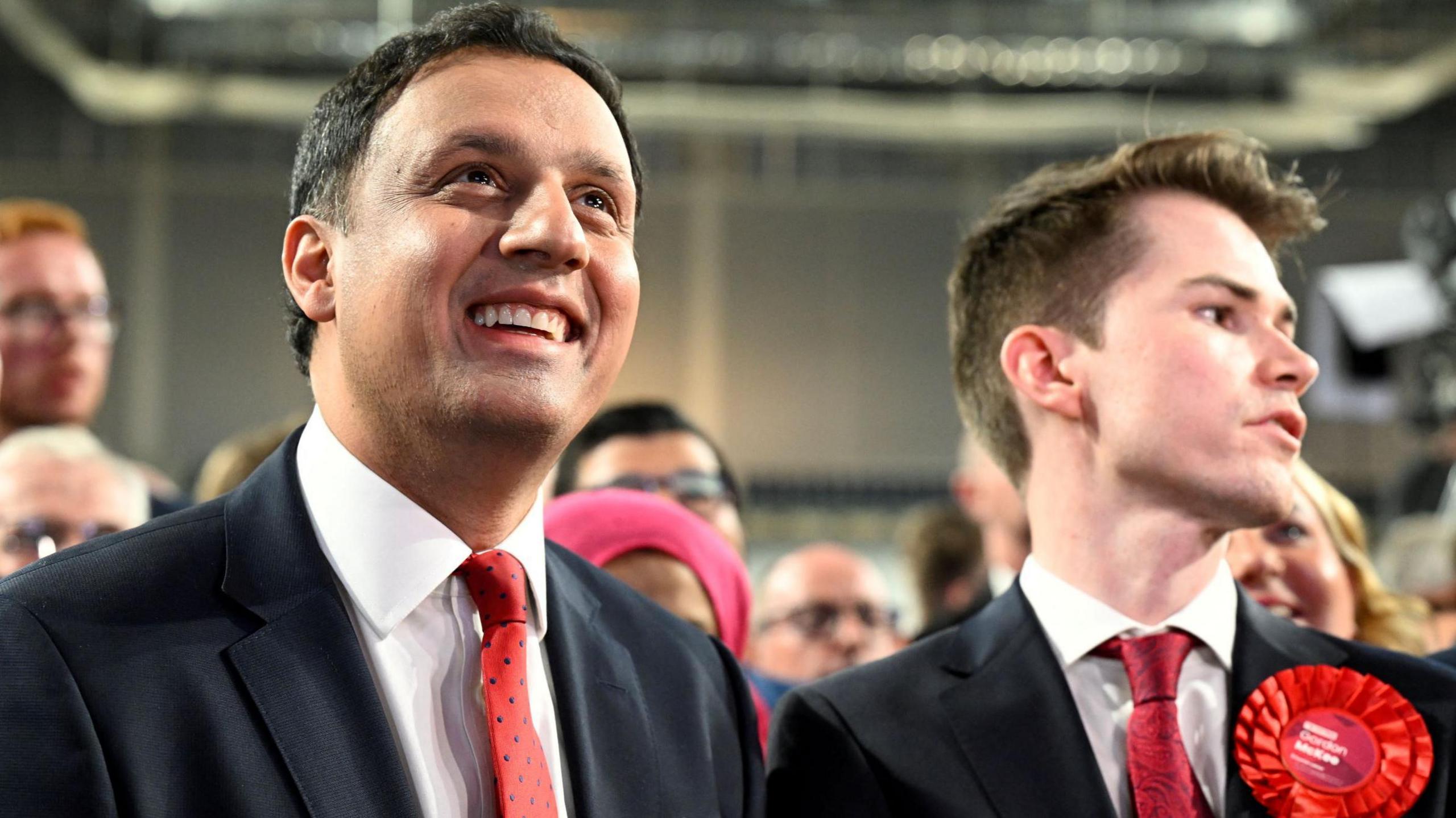 Anas Sarwar on left of screen in black suit, red tie, white shirt. Beside him is a young man in black suit, white shirt, red tie, with a Labour rosette. Sarwar is very happy looking. He appears to be looking at something in the distance. They are in an industrial-looking building.