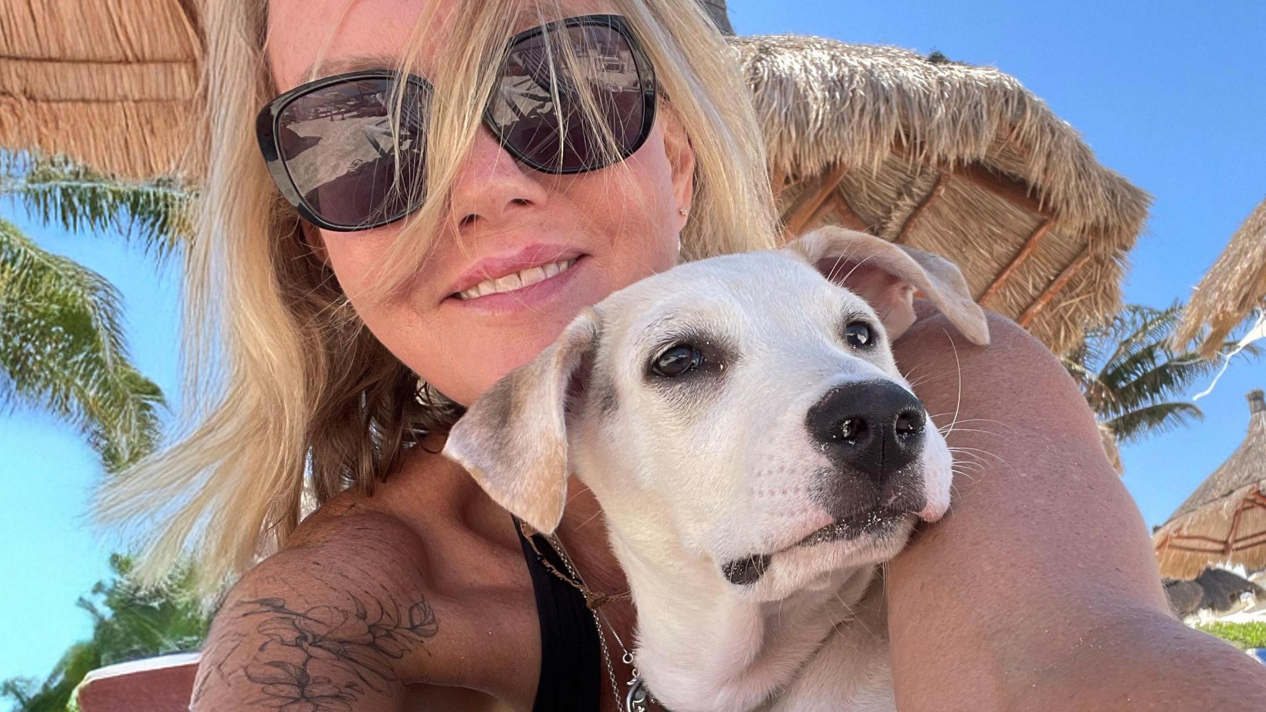 Kathryn Grace is holding a pale dog in her arms. It looks like she is on a beach and has long blond hair and sunglasses on
