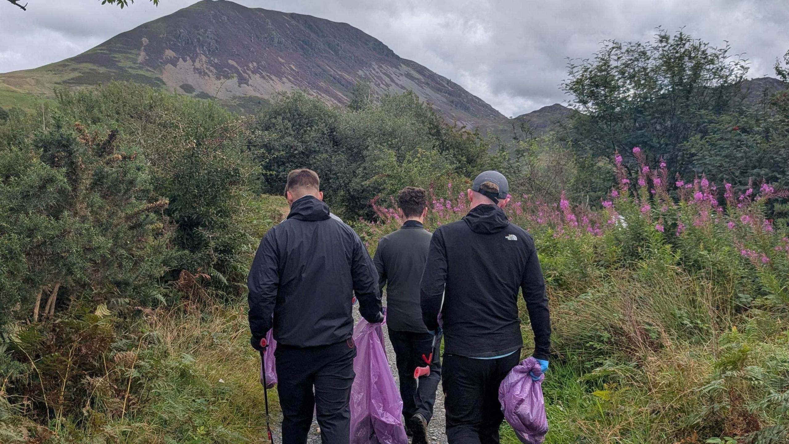 Three young men in dark clothing pictured from the back walking down a path at Ennerdale. They are carrying purple bin bags.