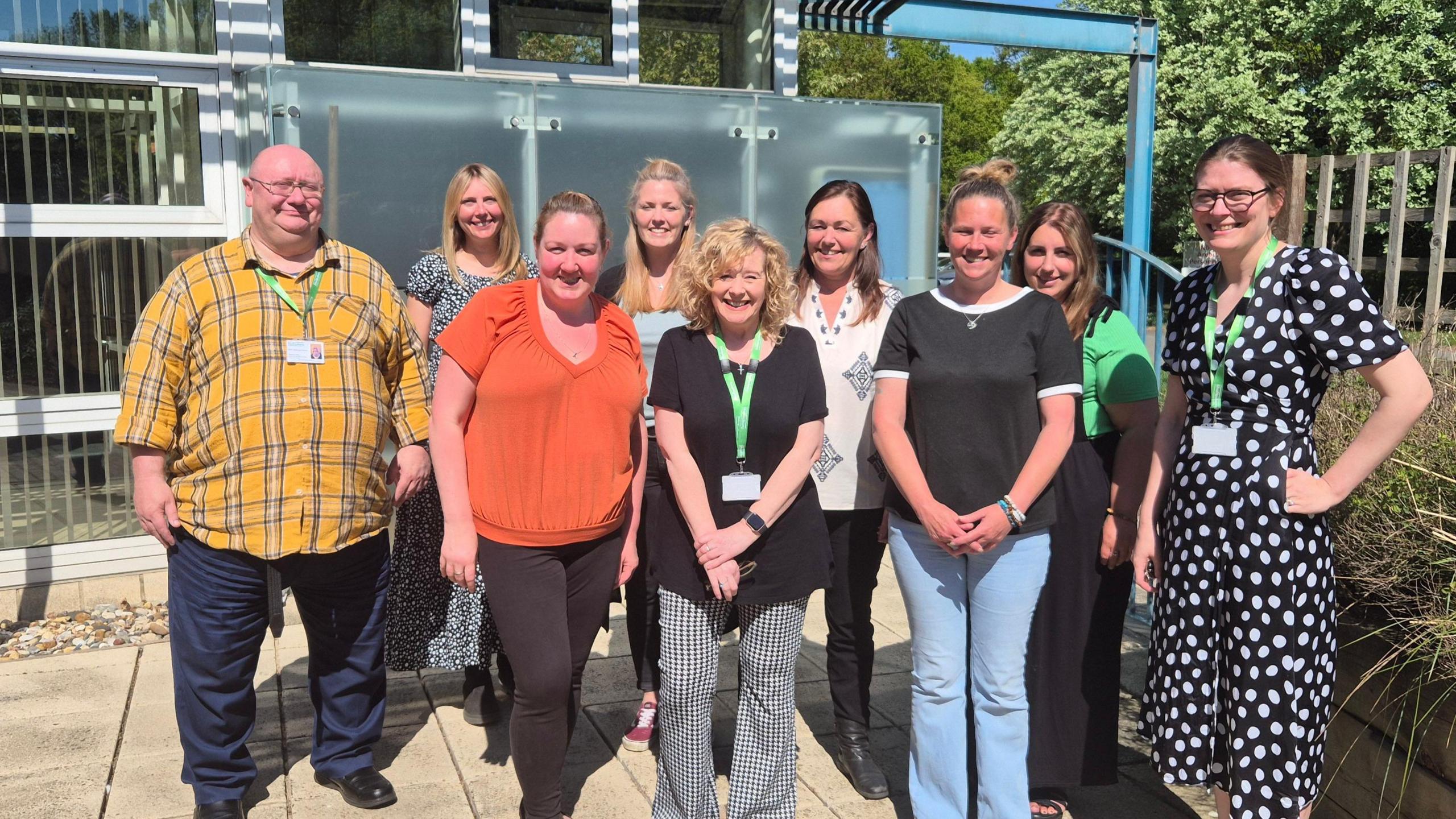 The Shaftesbury Suffolk Memory and Dementia Support team stand in a group outside and smile at the camera. Nickie Smith is pictured at the front of the group. She has curly short blonde hair and is wearing a black top with black and white chequered trousers.