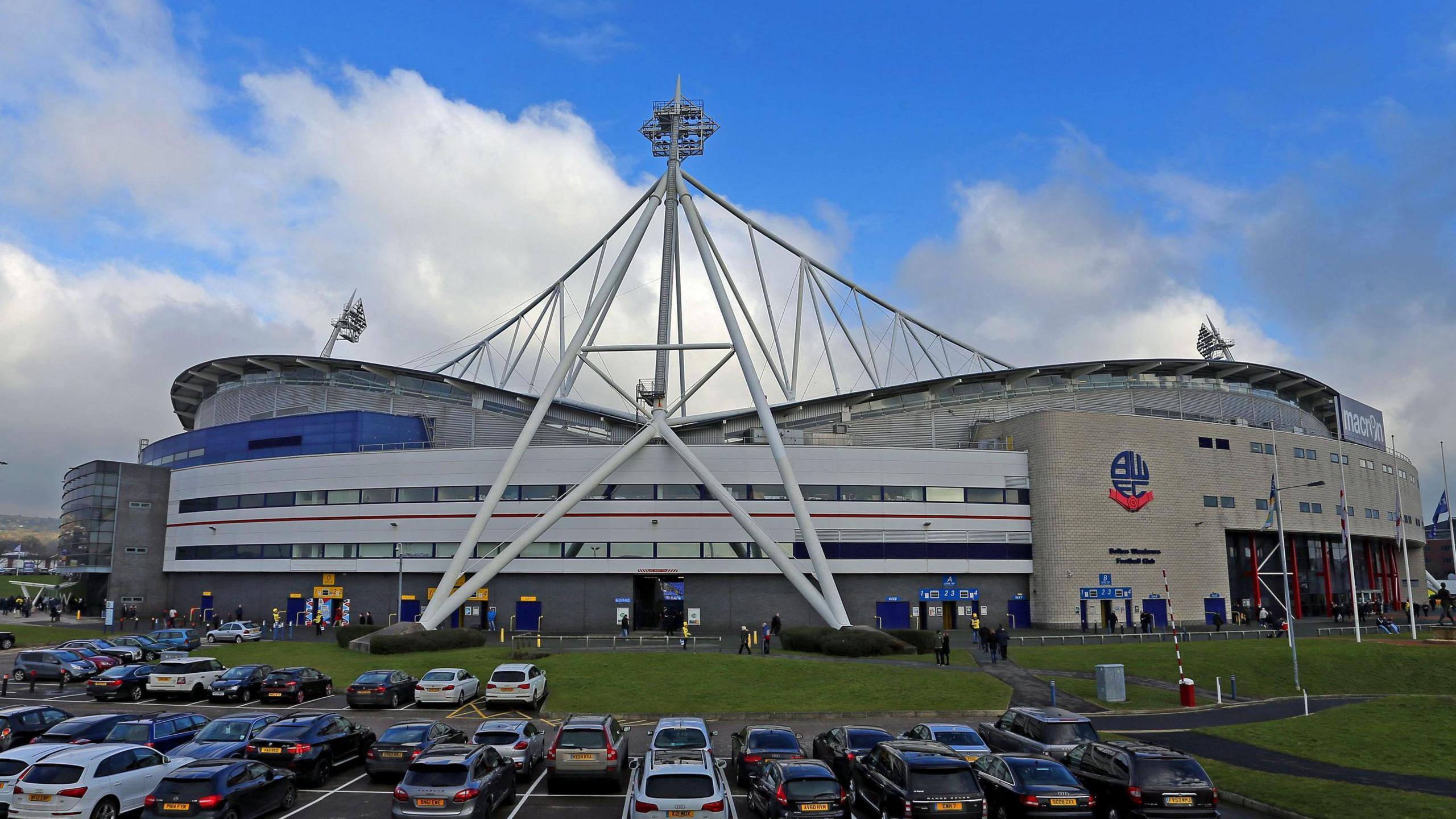 A football stadium viewed from a car park with a large white sail-like structure over a curved stand. The Bolton Wanderers club crest is visible on the side of the building