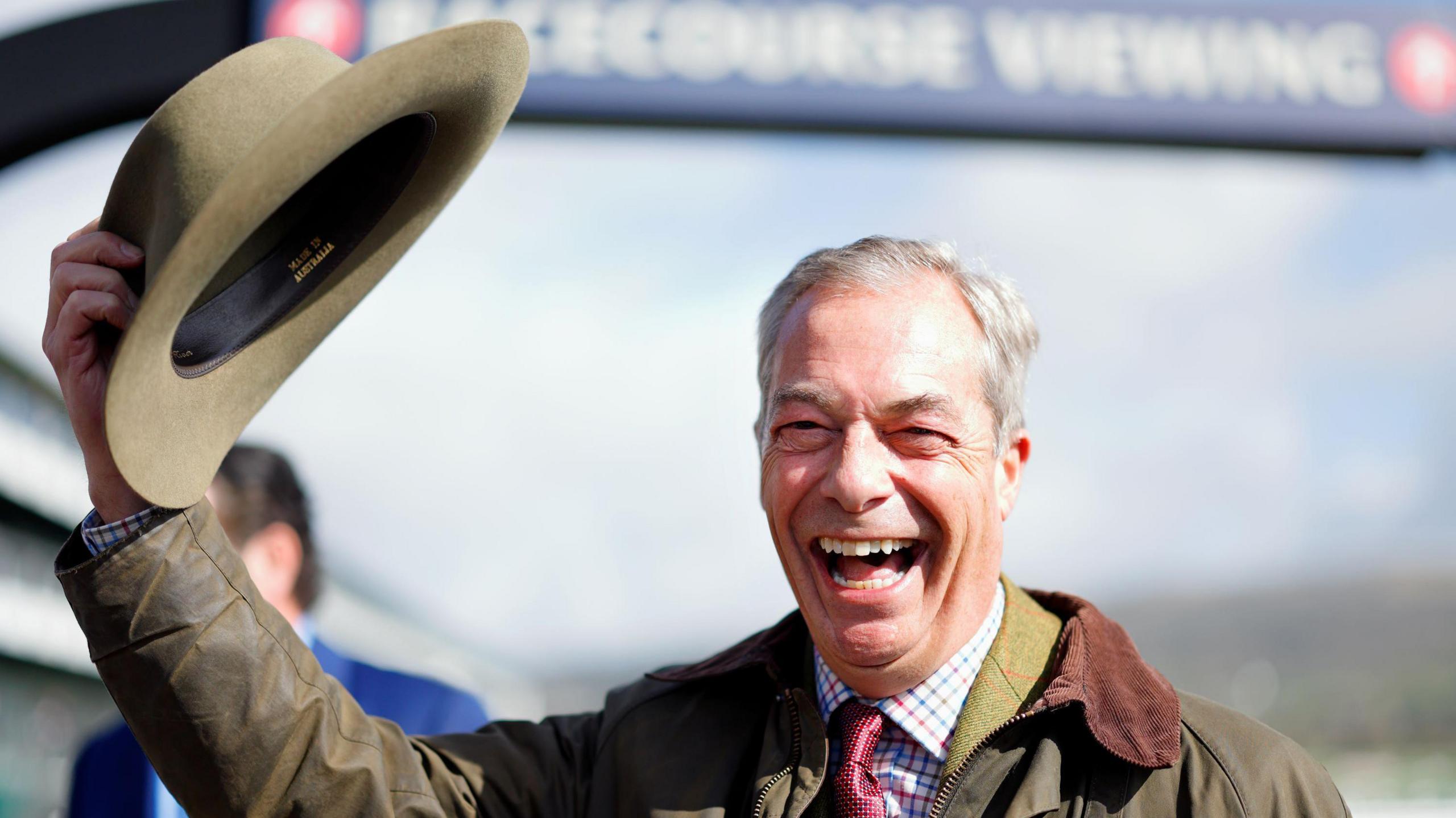 Reform UK leader Nigel Farage in a wax jacket laughs as he lifts up his hat in front of a racecourse sign