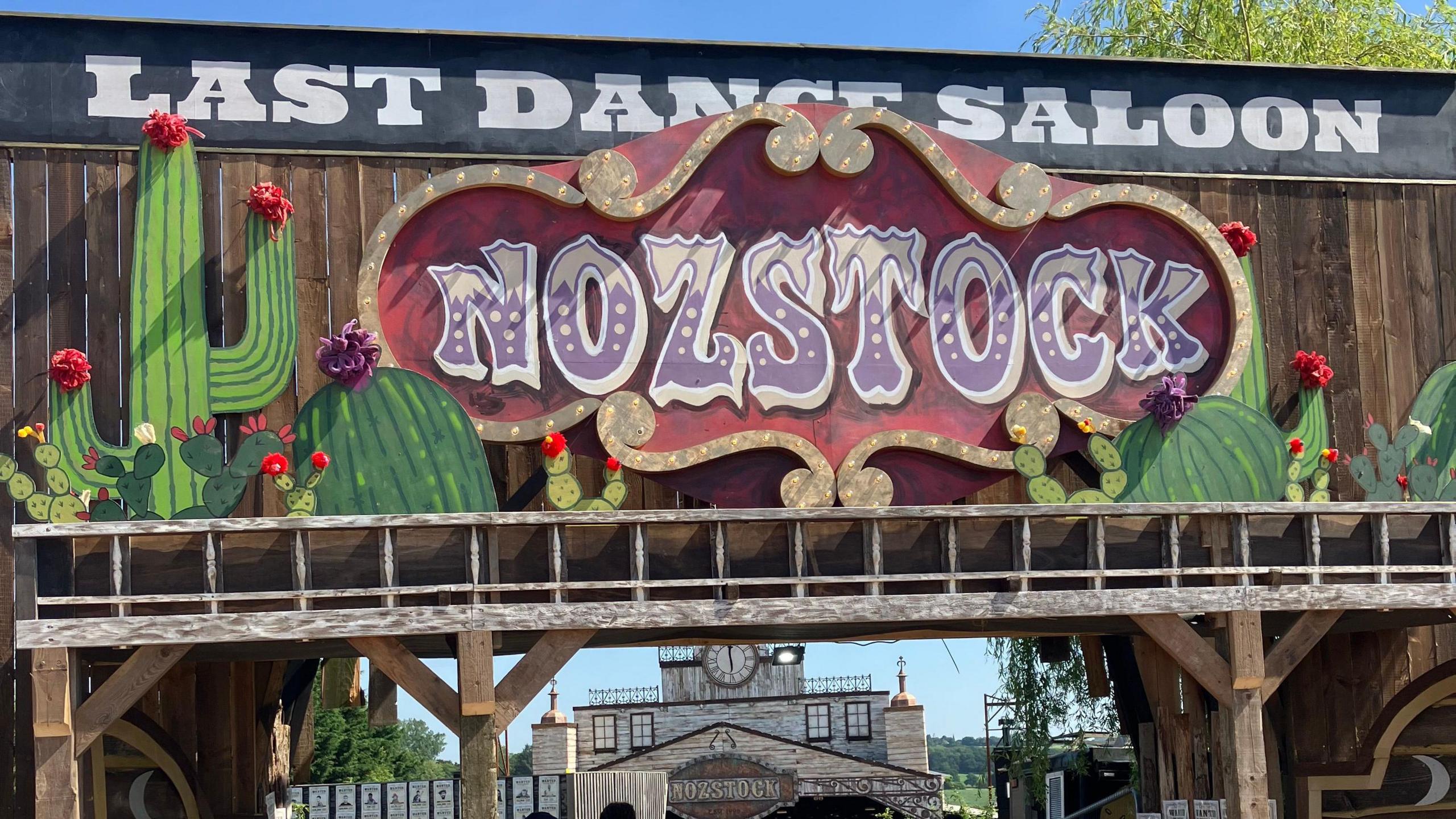 A brown wooden backdrop has a sign reading "Nozstock" in purple and white writing, surrounded by red and gold. There are green cacti painted next to it, and a sign in black and white that says "last dance saloon"