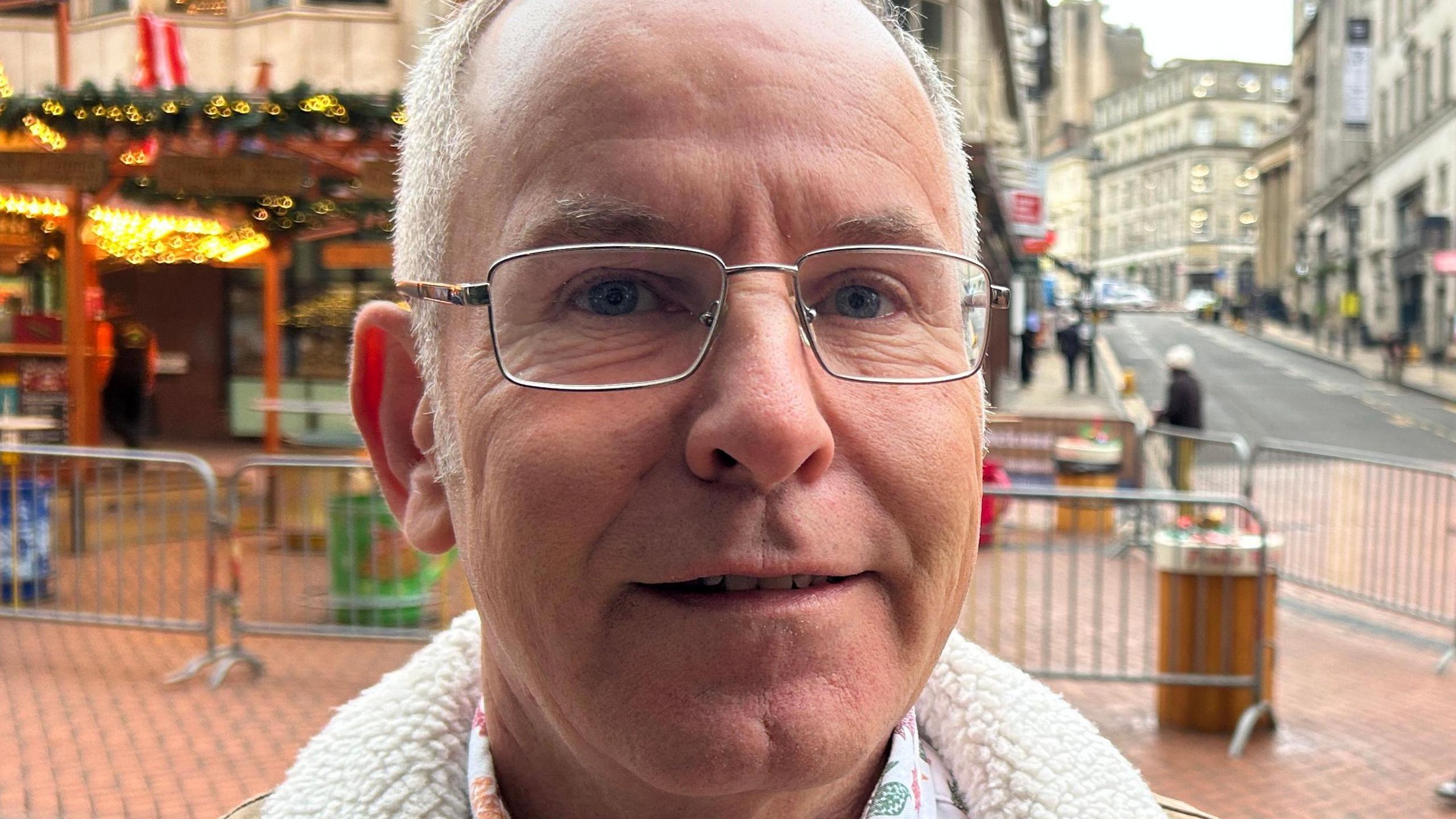 Nick Vaughan is wearing glasses. He has grey hair and is smiling. He is stood in front of the Birmingham Christmas market. 