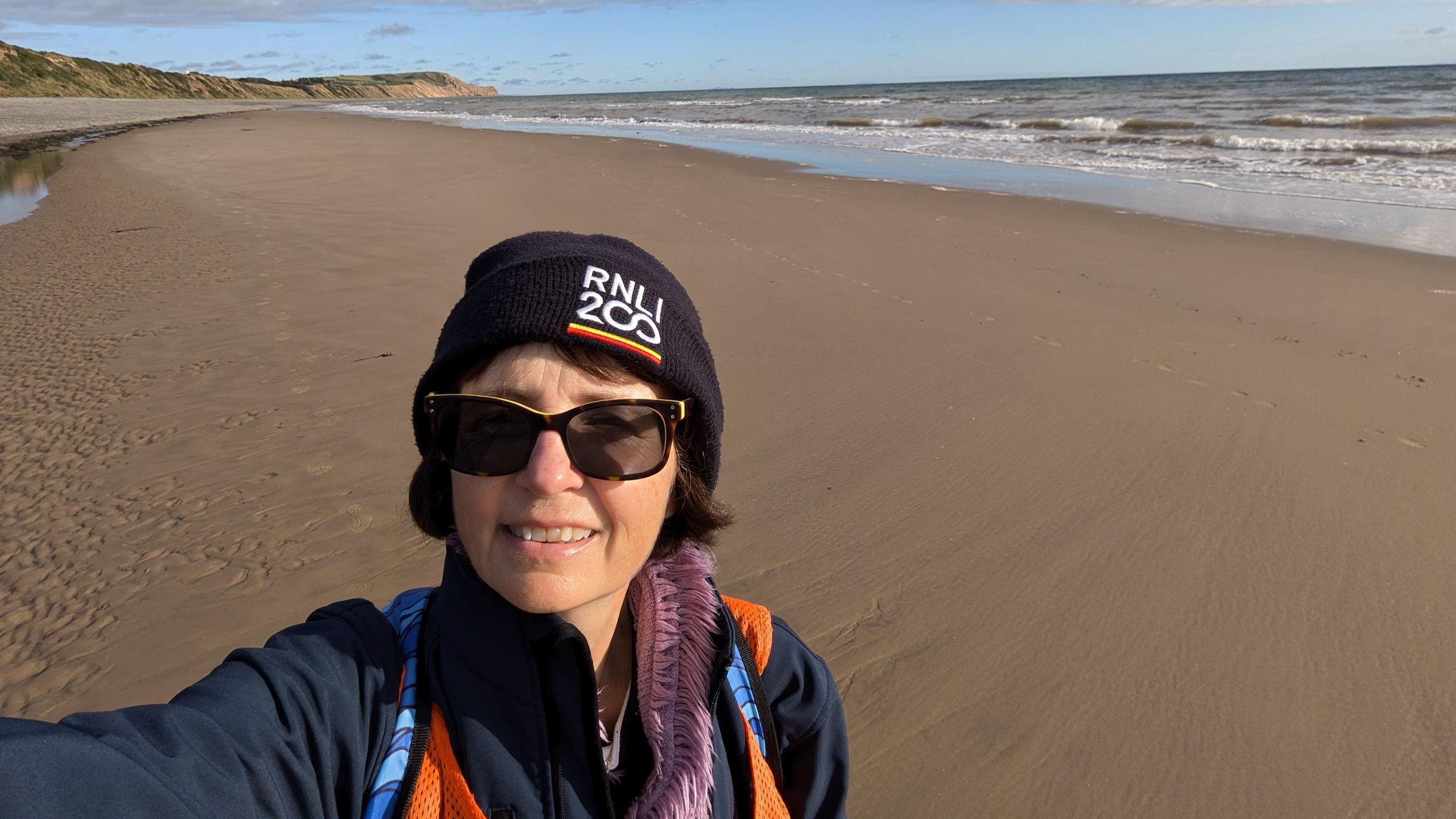 Anjie Rook holding with her arm raised up taking a selfie on a sandy beach where the tide is out. She is wearing sunglasses and a navy blue hat with RNLI 200 on the front.