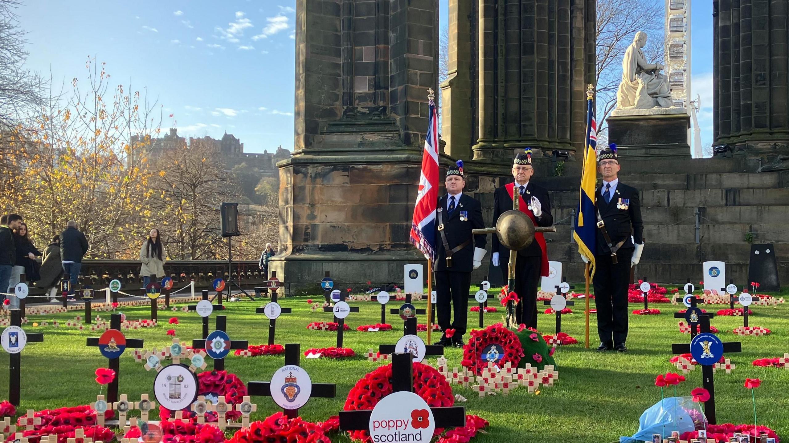 Three veterans stand with a rustic cross adorned with a WWI helmet in front of the Scott Monument in Edinburgh. There are poppy wreaths and black crosses mounted in the grass. Edinburgh Castle sits in the distance.