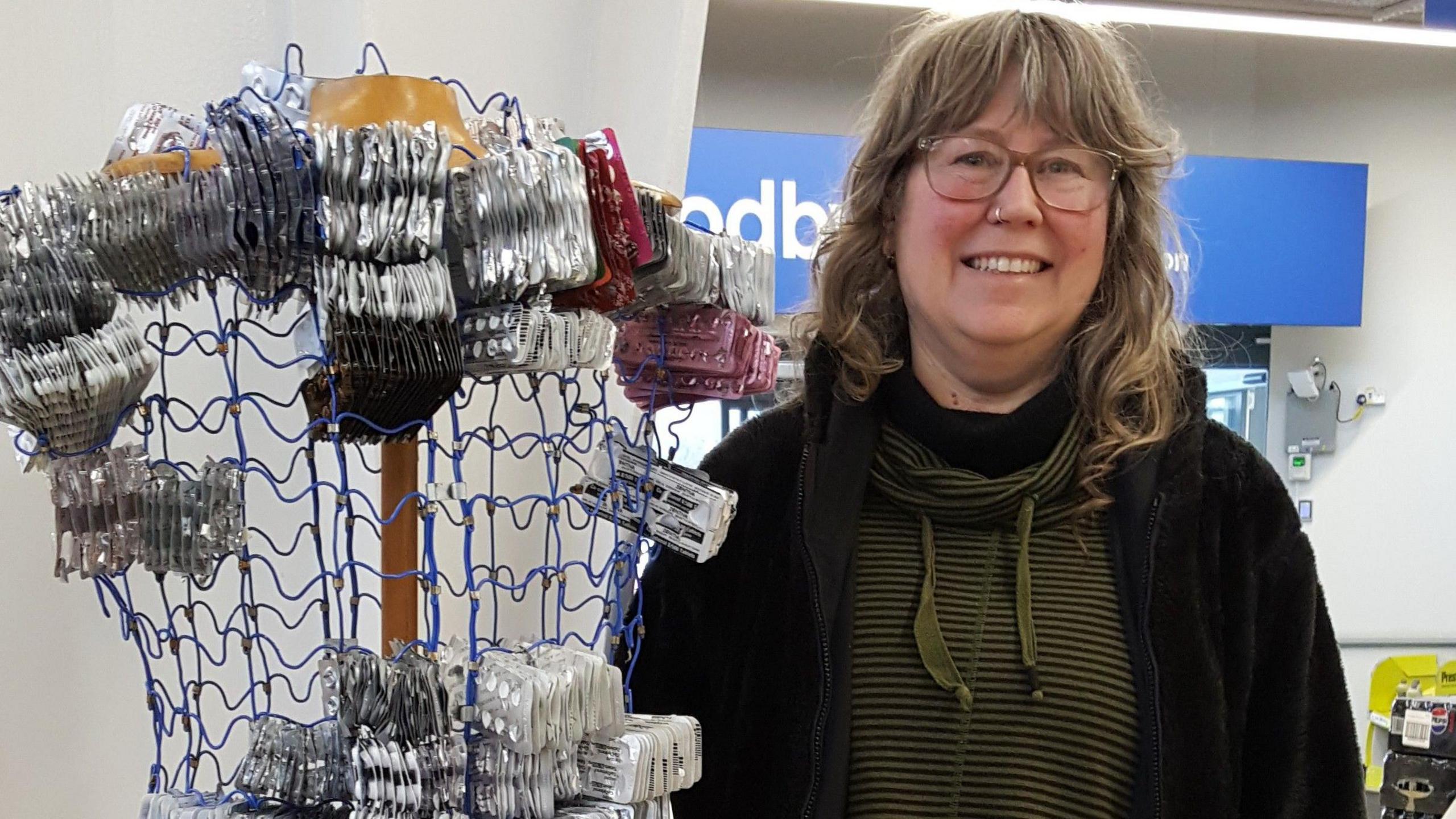 Miranda Goudge stands next to an artwork shaped like a dressmaker's dummy, which is made of wire and is slowly being filled with used blister packs.