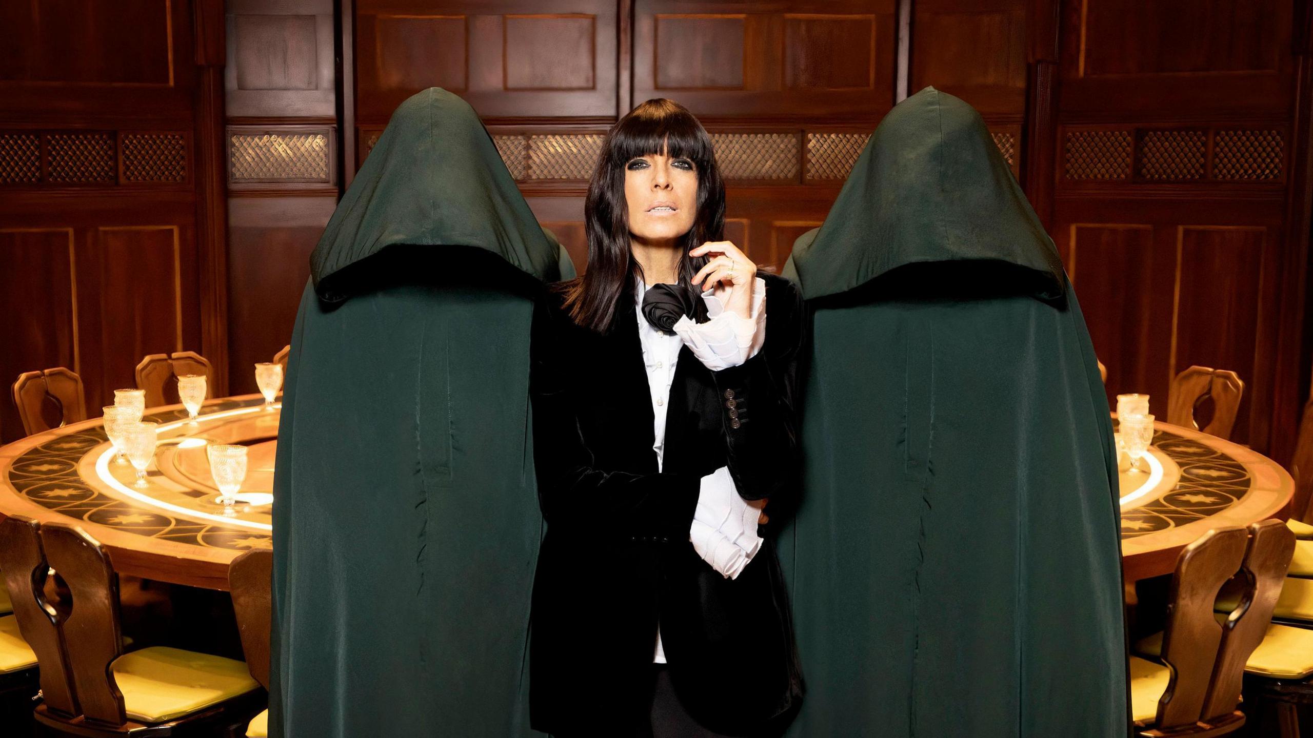 Claudia Winkleman flanked by two cloaked and hooded figures standing in front of the round table in a wood-panelled room. 