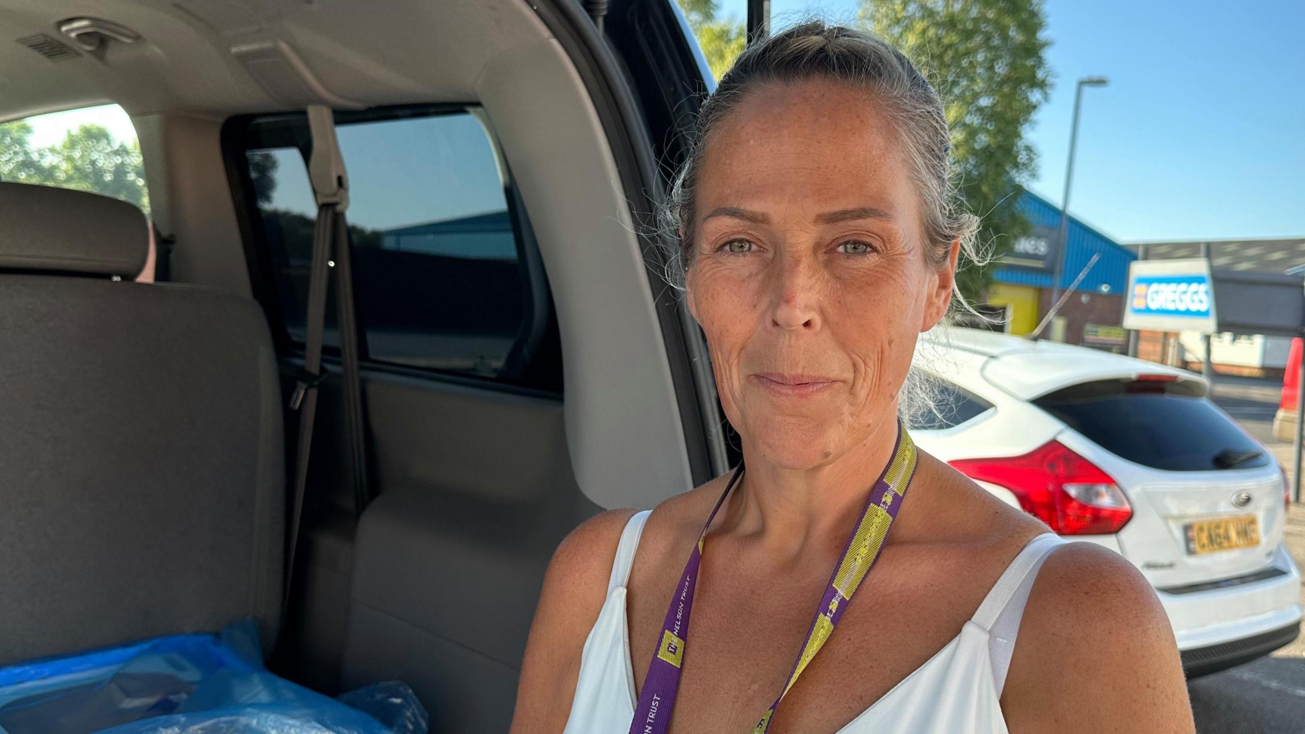 A woman in a white vest standing at the open boot of a van in a car park looking at the camera