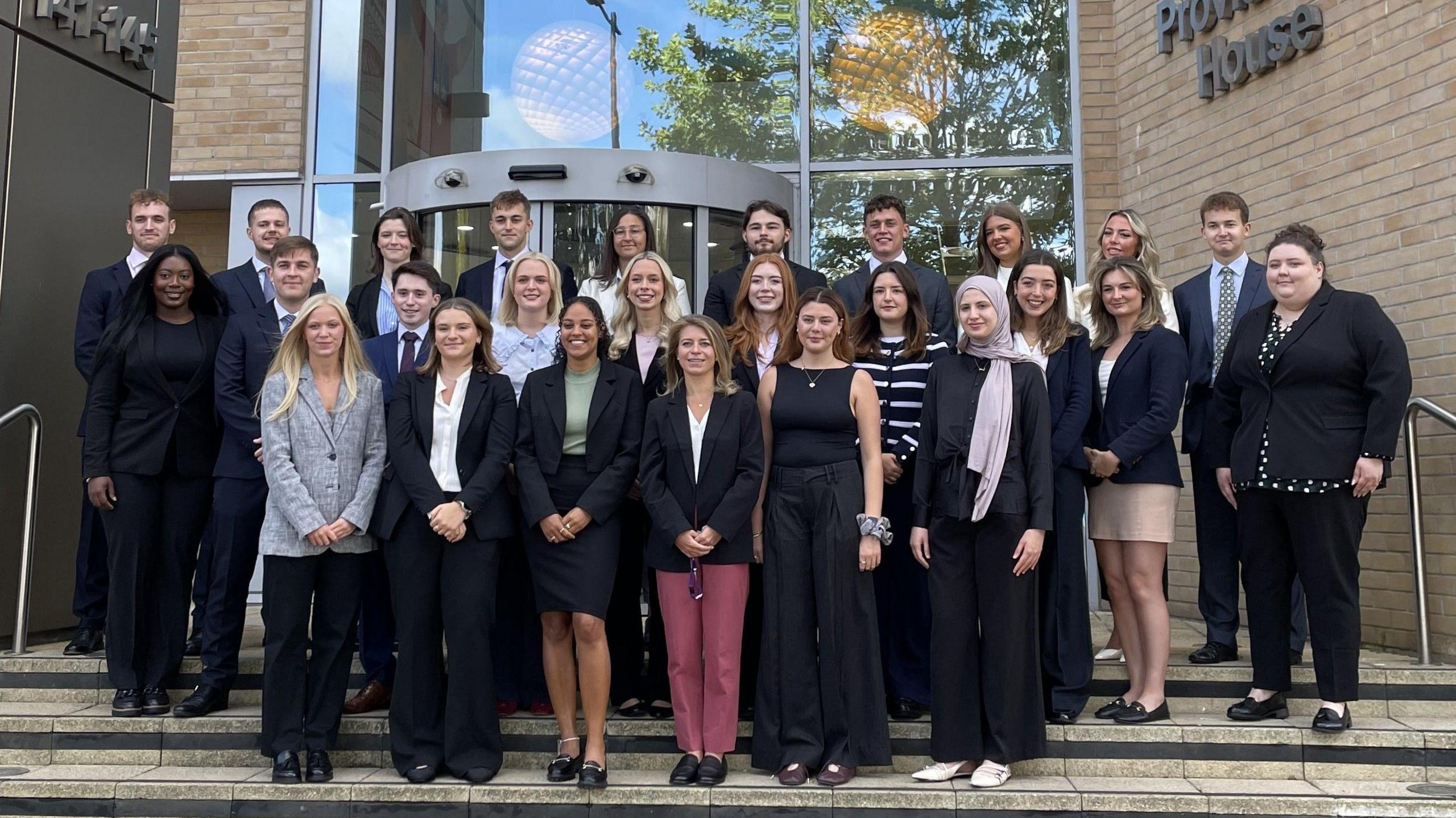 A group of about 30 trainee solicitors, the majority of them women, stand wearing smart office clothes on the steps of a building. Behind them is a revolving glass entrance door with large windows above and to the side of it