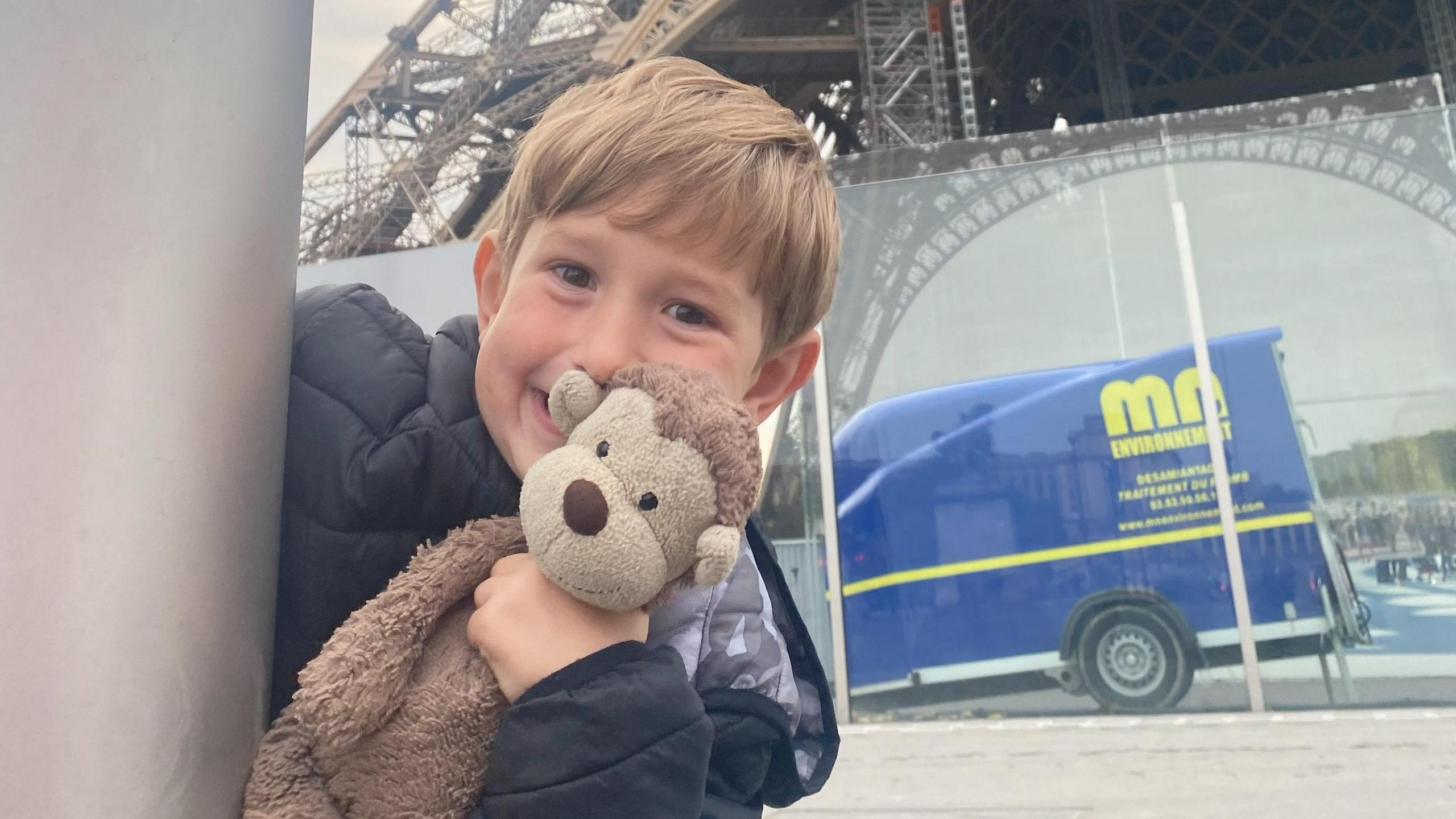 A young boy peeking round the corner of a grey post smiling in a cheeky way and holding a brown monkey teddy up to his cheek. He has dark blond hair and a fringe and is wearing a black coat. The bottom of the Eiffel Tower is in the background, as is a reflection of a blue van.