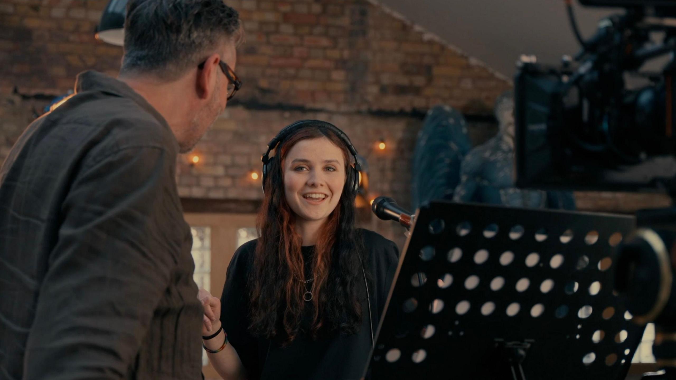 Grace Cope stands behind a microphone and camera. She is wearing wearing headphones and smiling at a man in the foreground who is side-on to the camera. A music stand can also be seen, and there is a brick wall behind her.