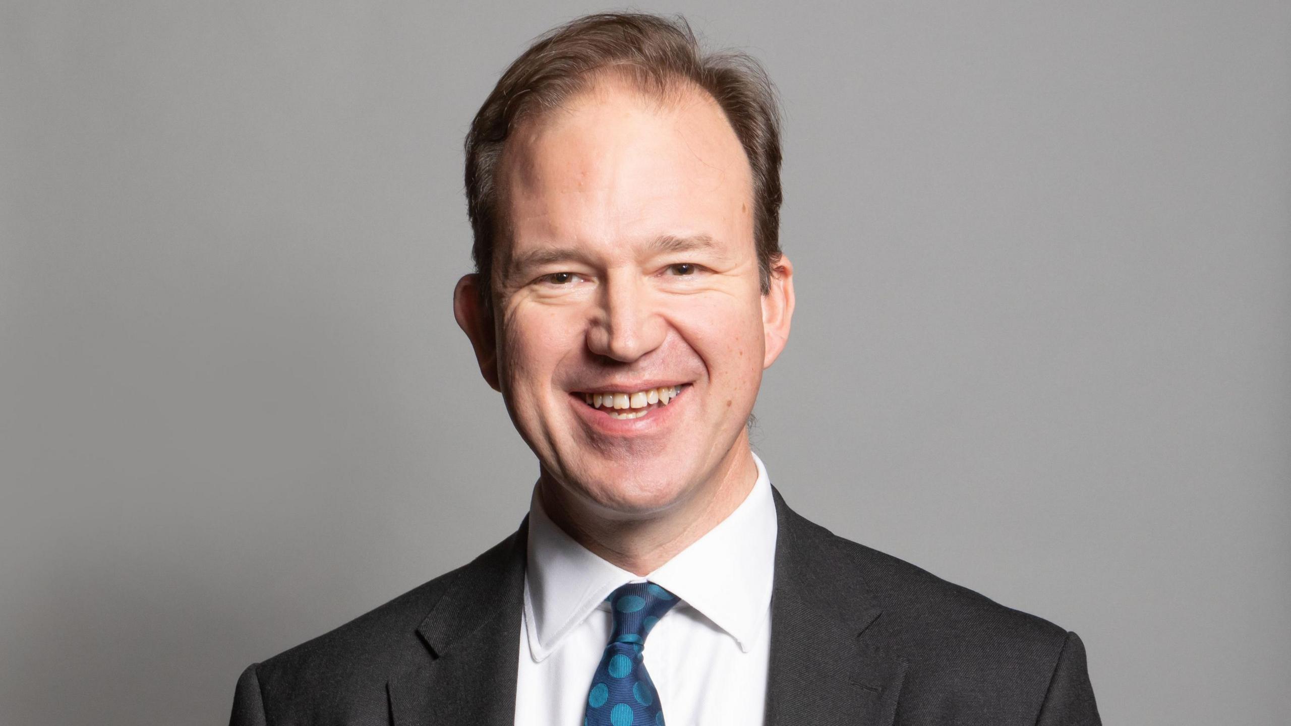 A portrait photograph of MP Jesse Norman. He is wearing a grey suit jacket and white shirt with a blue spotty tie. He is stood against a grey backdrop.