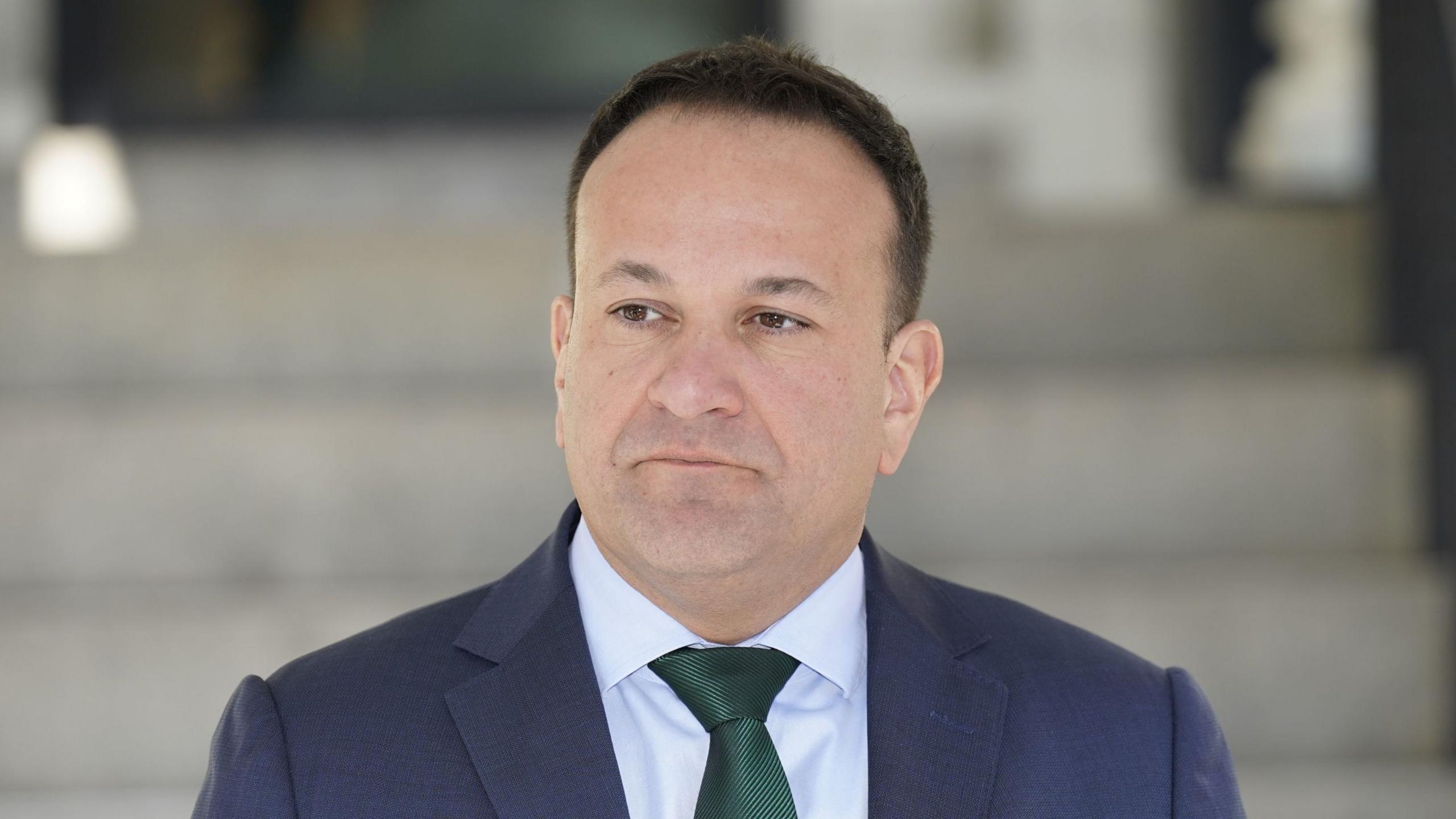 Leo Varadkar has short dark hair. He is looking off to the side of the camera. He is wearing a blue suit jackers, a white shirt and a green tie.
