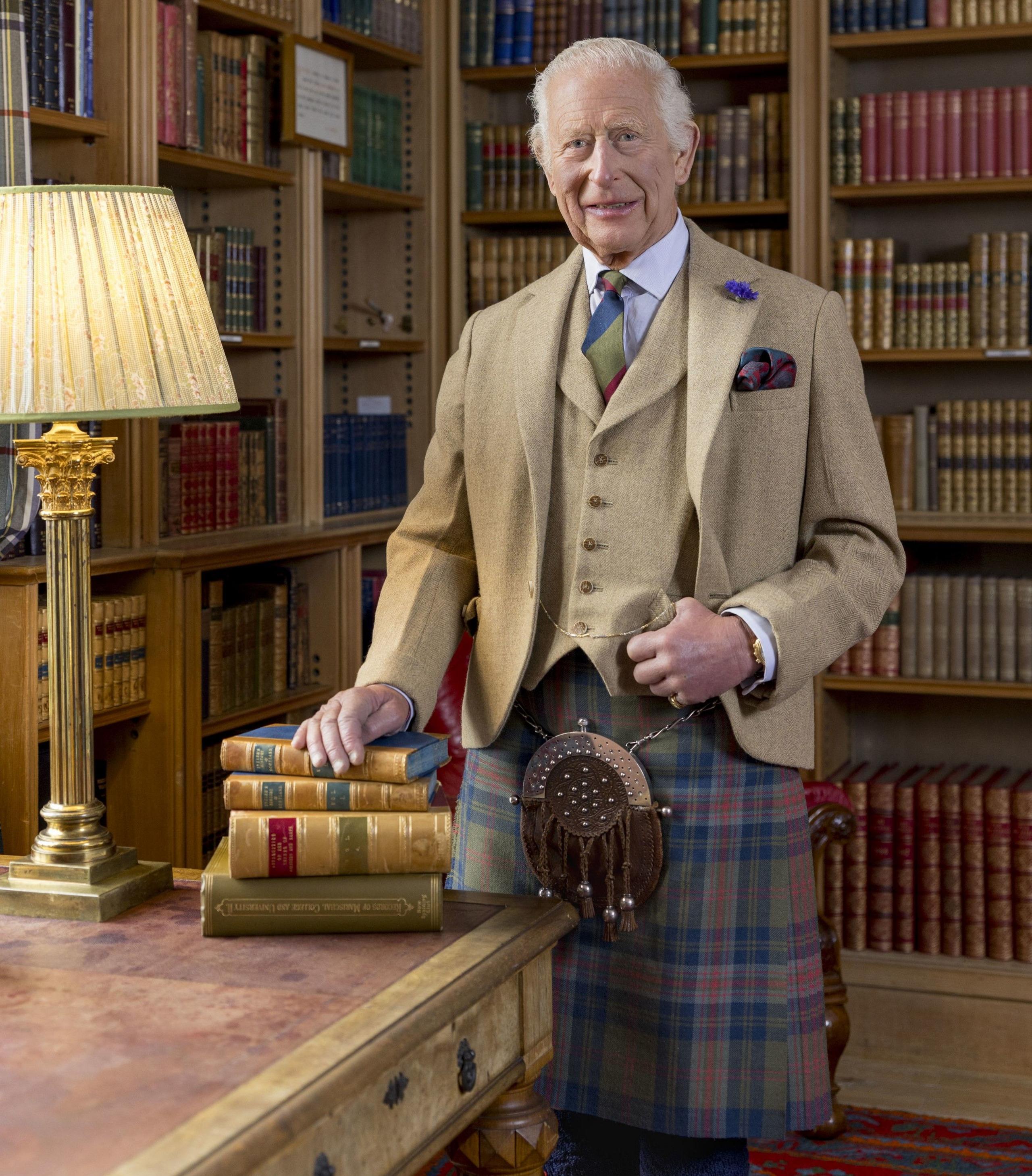 King Charles is wearing a beige jacket and waistcoat. He stands in front of a collection of books in a library and is smiling at the camera