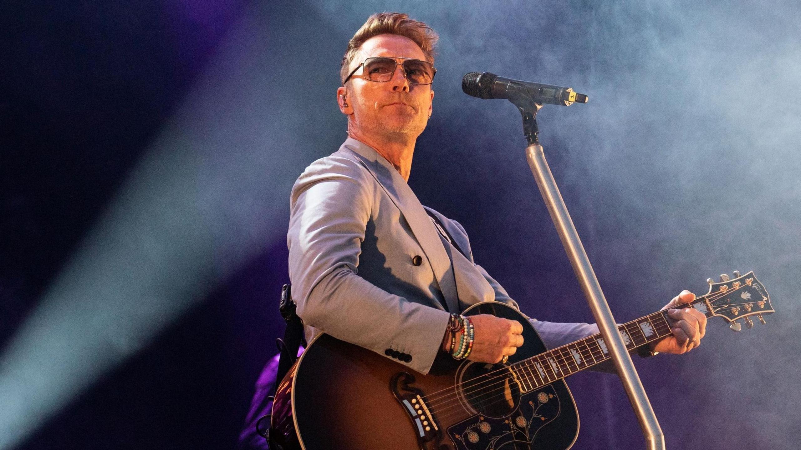 Ronan Keating performing onstage. He's wearing a suit and holding a guitar. A mic is in front of him. Smoke is in the air and a spotlight is on him. He's wearing glasses.