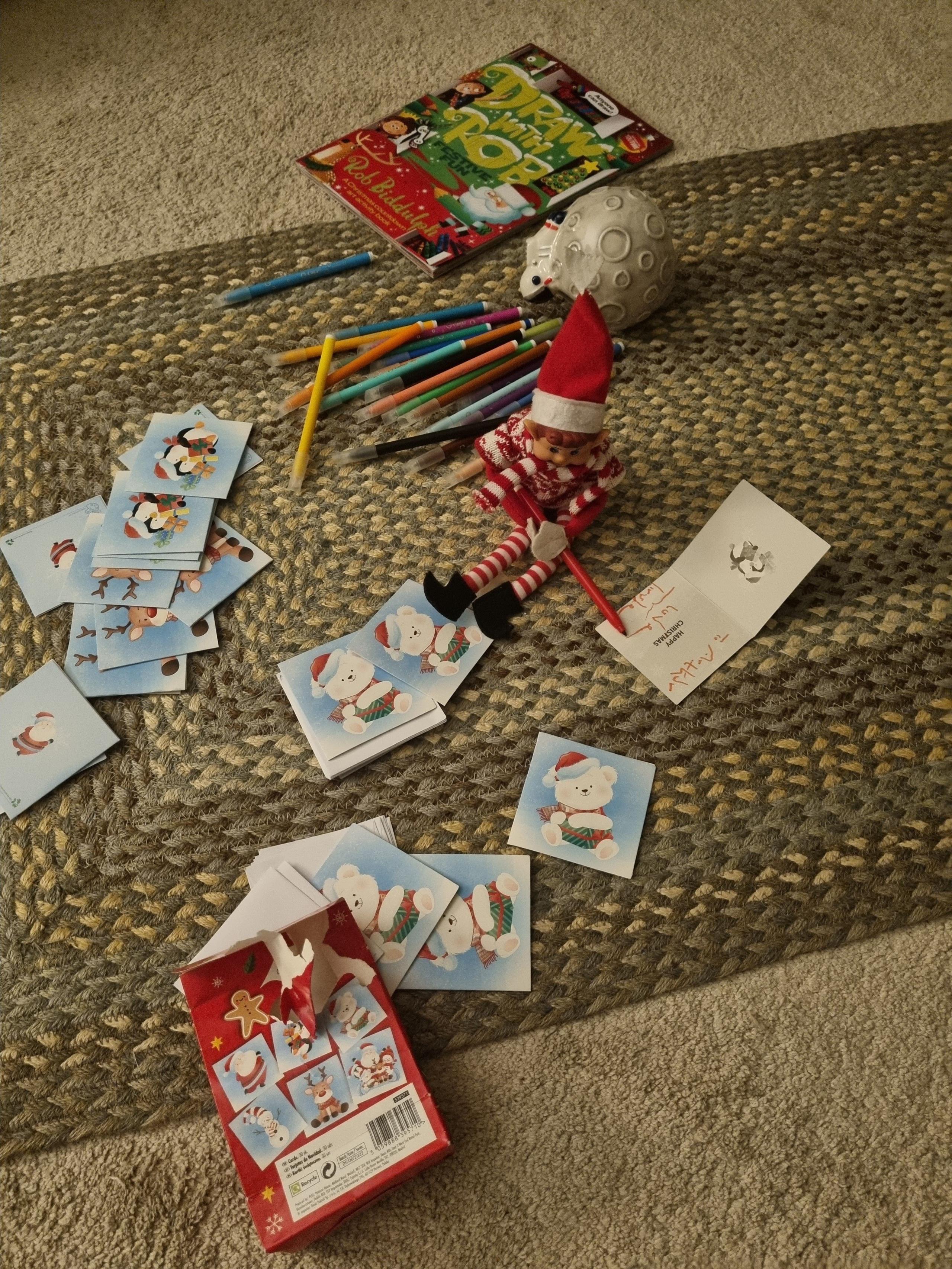 An elf sat on the floor holding a pen with Christmas cards all around it.