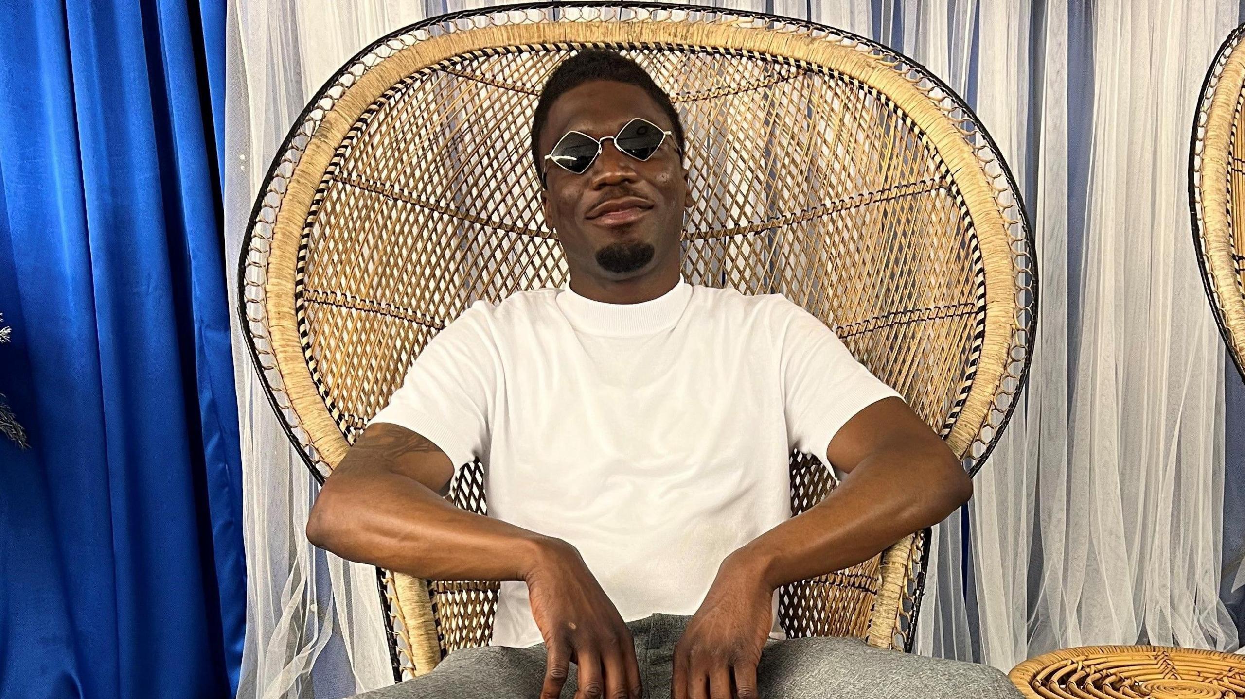 Alex Mamwa wearing grey jeans and a white t-shirt. He is sitting in a large rounded wicker chair, wearing black diamond shaped sunglasses and smiling at the camera. He looks relaxed with his elbows resting on the armrests. 