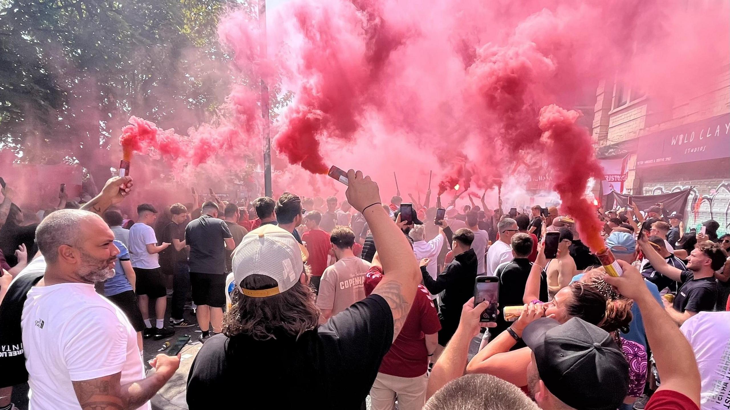 Three men, facing away from the camera, hold up red smoke flares as part of the Bristol City flag day event. Beyond them dozens of other fans are doing the same, and a large cloud of red smoke is hanging over the street