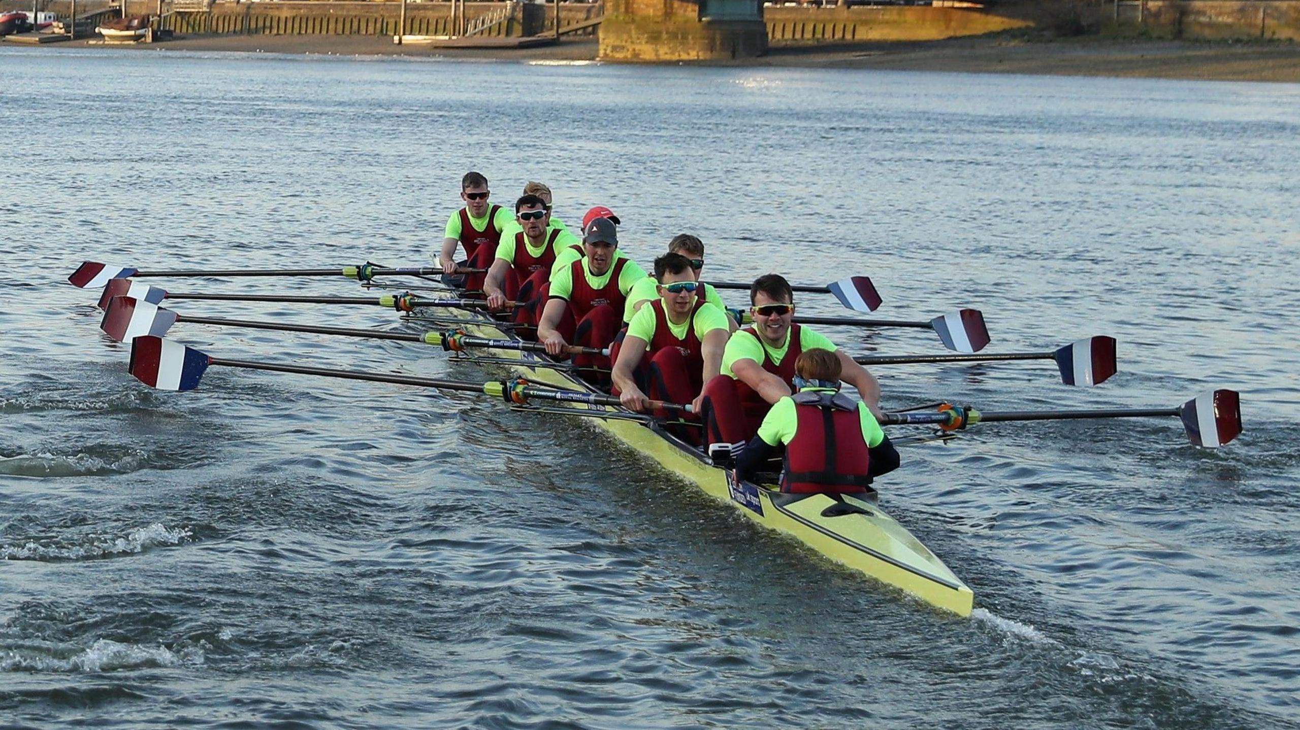 Nine members of Oxford Brookes University Boat Club, all wearing burgundy, row on a stretch of water. Their ores are stripes red, white and blue. Their rowing boat is yellow. Most members are male and are wearing sunglasses.