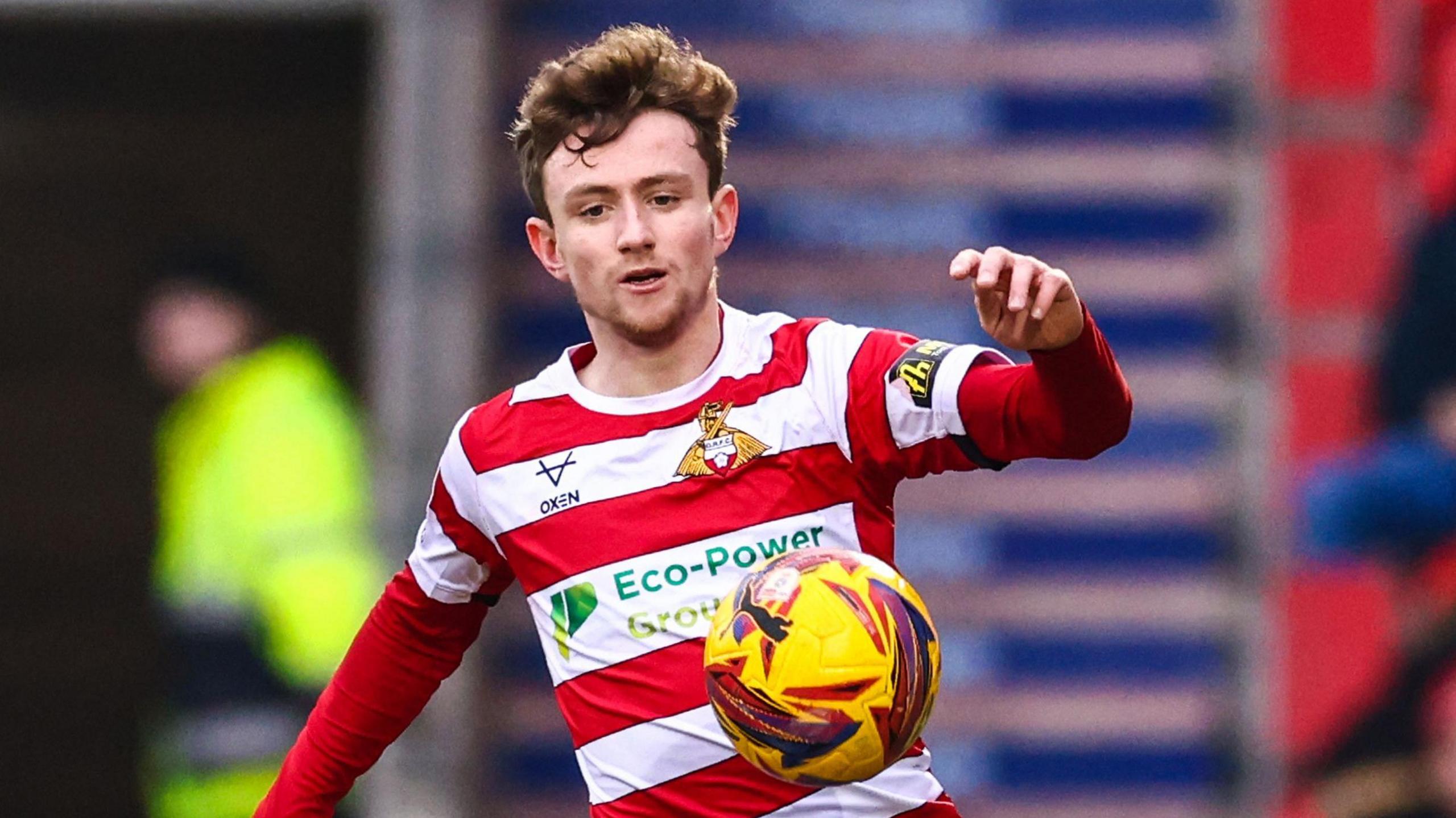 James Maxwell is wearing a red and white hooped Doncaster shirt and is trying to control the ball as he runs forward.
