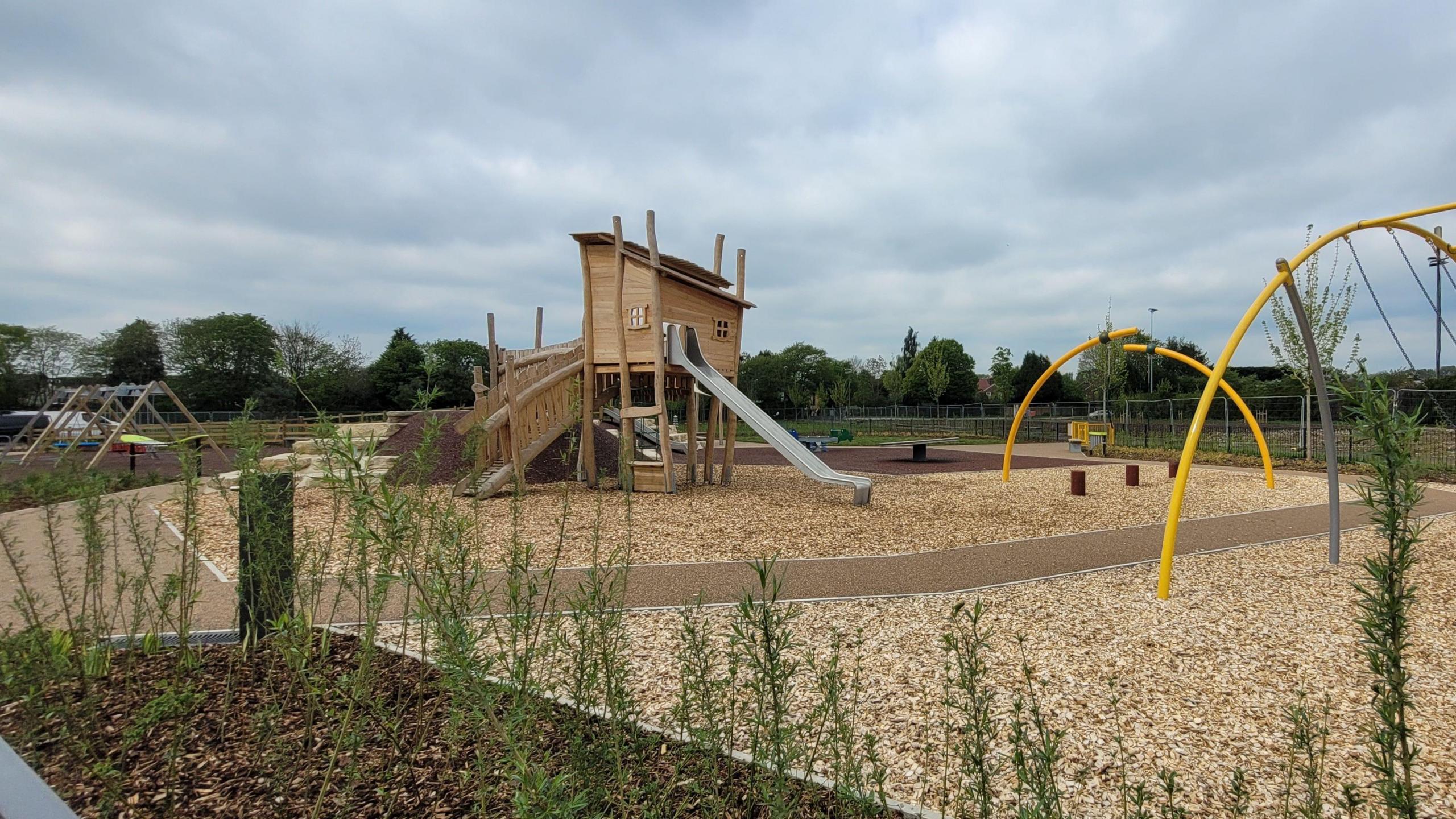 A wooden play area with a slope leading up to one side of a building on long legs and a metal slide coming out another side. The floor is woodchipped and there are yellow metal bar structures with swings on them.