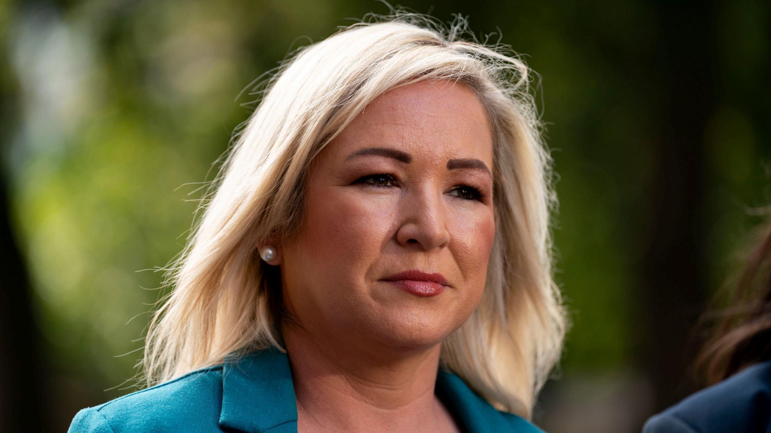 A woman with shoulder length blonde hair looks off to the side of the camera lens. She is wearing a teal coloured formal blazer and pearl earrings. The background is blurred but you can see the outline of foliage. 