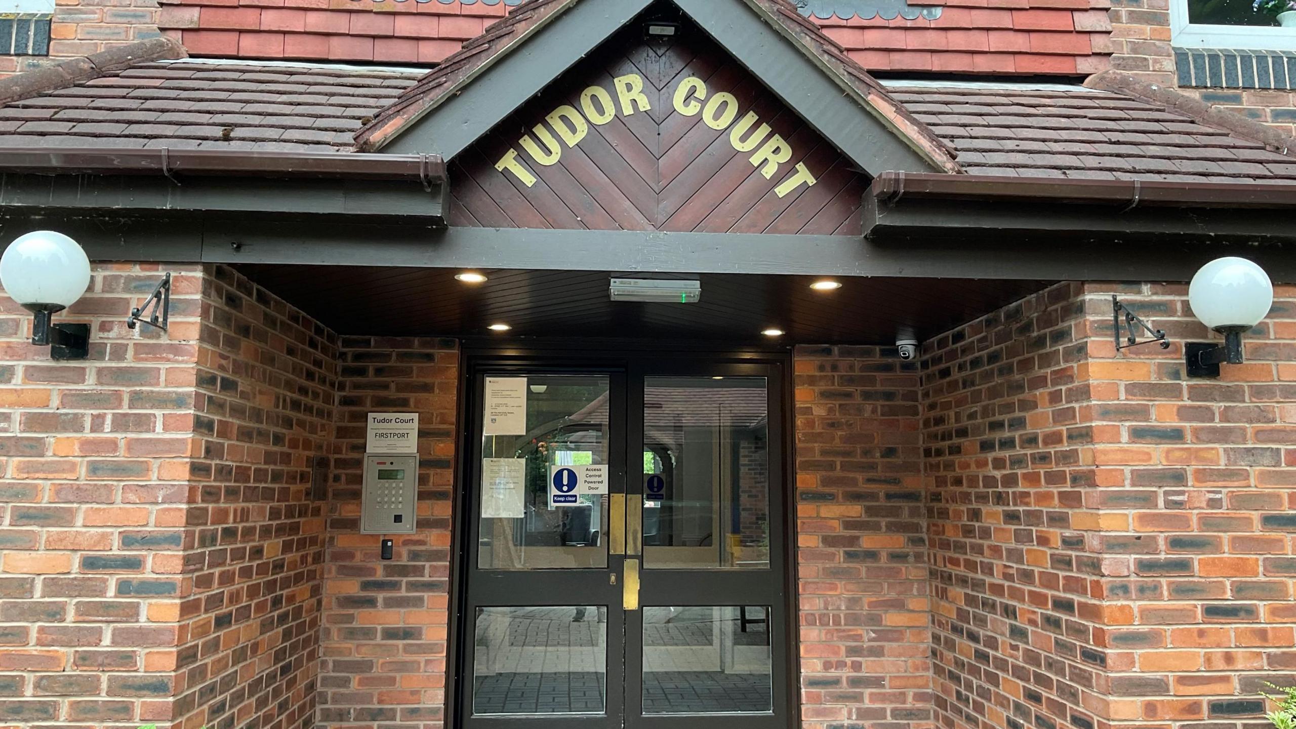 The outside of Tudor Court. It shows a brick entrance, glass doors and has the sign Tudor Court in gold writing at the top.