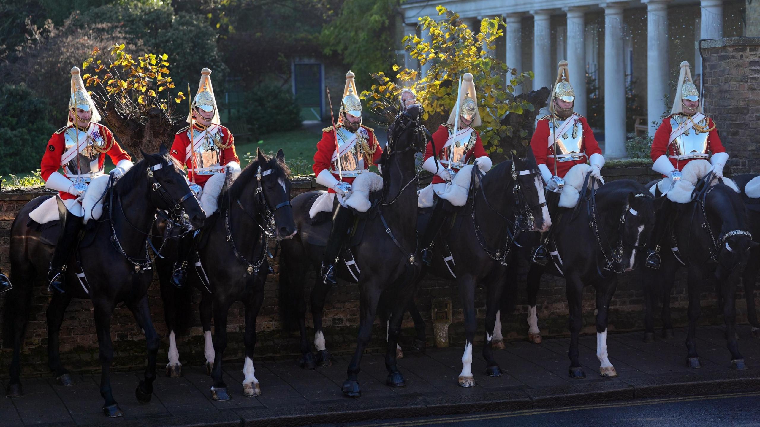 Burra të veshur me kostume të kuqe ceremoniale mbajnë shpata të ulura mbi tuba para një muri guri